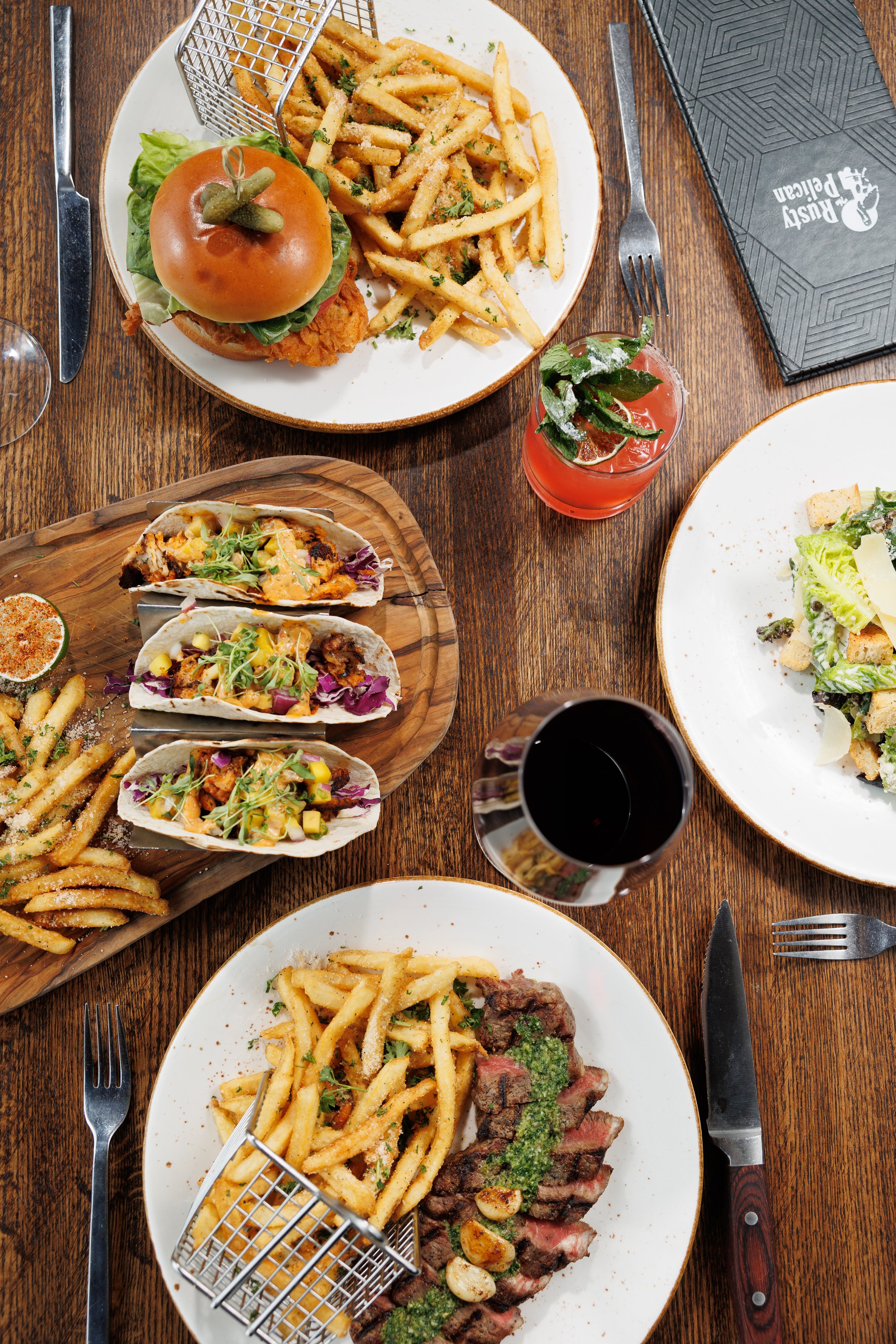 A table with various plates of food including French fries, tacos, a burger with lettuce and pickles, a grilled steak with garlic and herbs, and a drink garnished with mint and lime. There are utensils and a black disposable placemat with a restauran