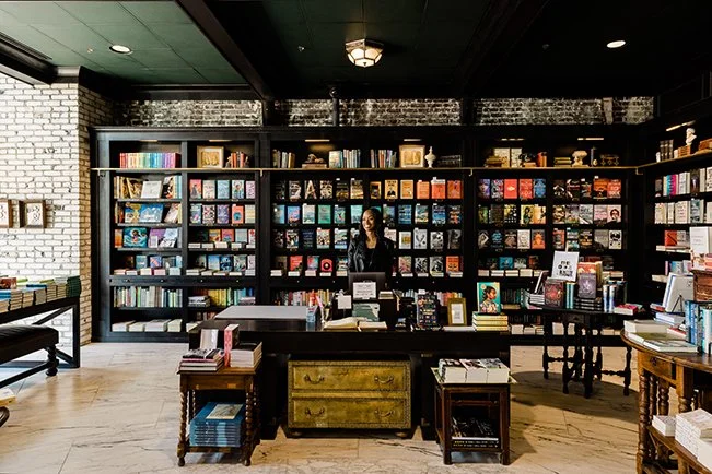 Interior of a bookstore with shelves filled with books, a woman standing behind a desk, and various books displayed on tables.