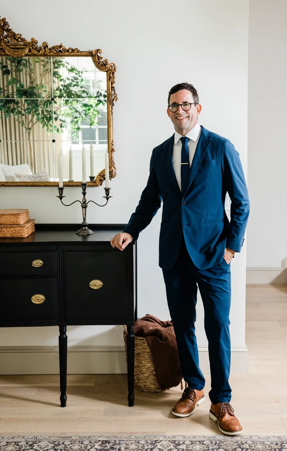 A man wearing a blue suit, white shirt, and dark blue tie standing and smiling in a well-lit room next to a black dresser with brass handles, a silver candelabrum with white candles, and a mirror reflecting a window with curtains and a plant inside.