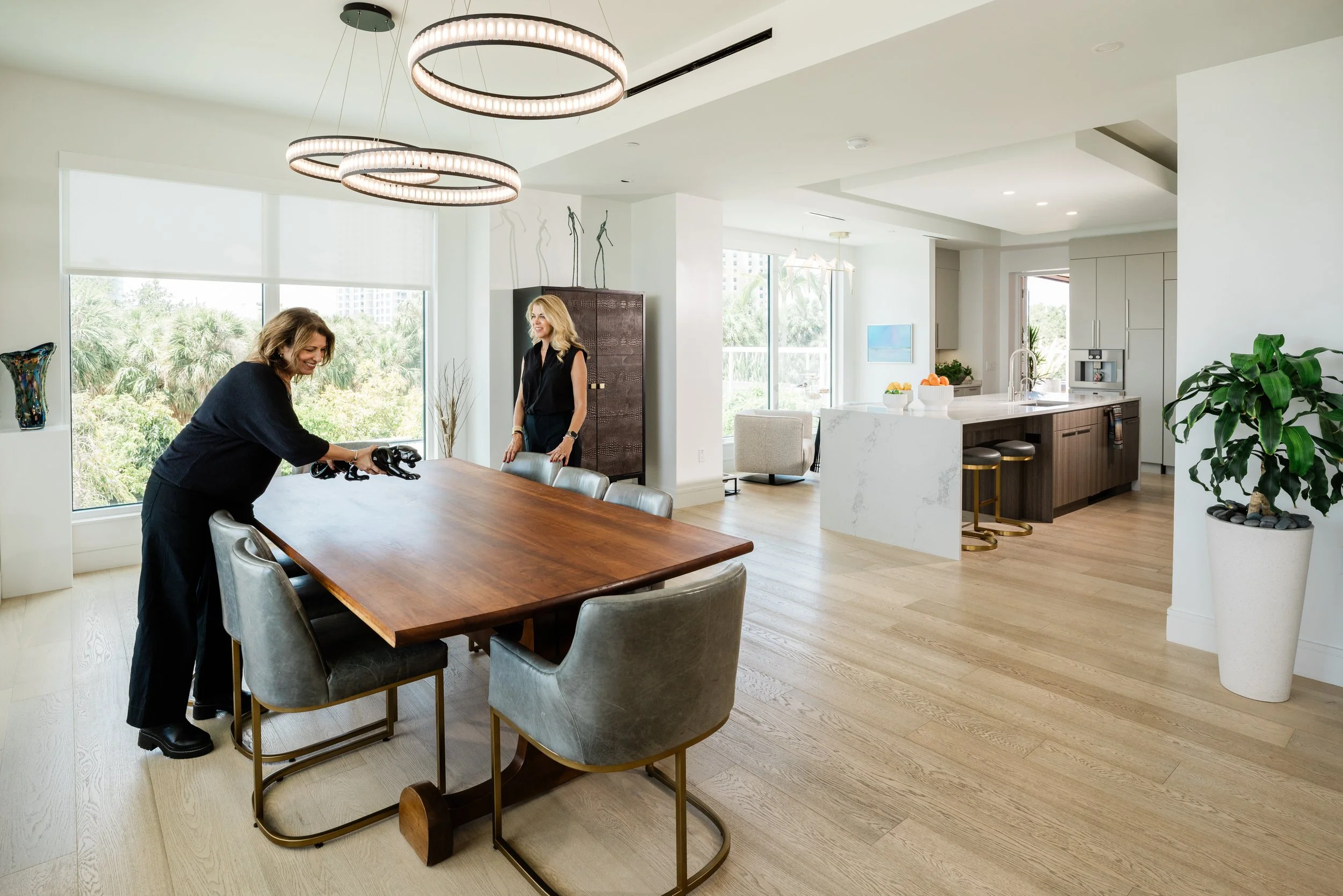 Two women are in a bright, modern kitchen and dining area. One woman is standing and smiling while the other woman is leaning over a wooden dining table, holding a black object. The kitchen has large windows showing greenery outside, a white countert