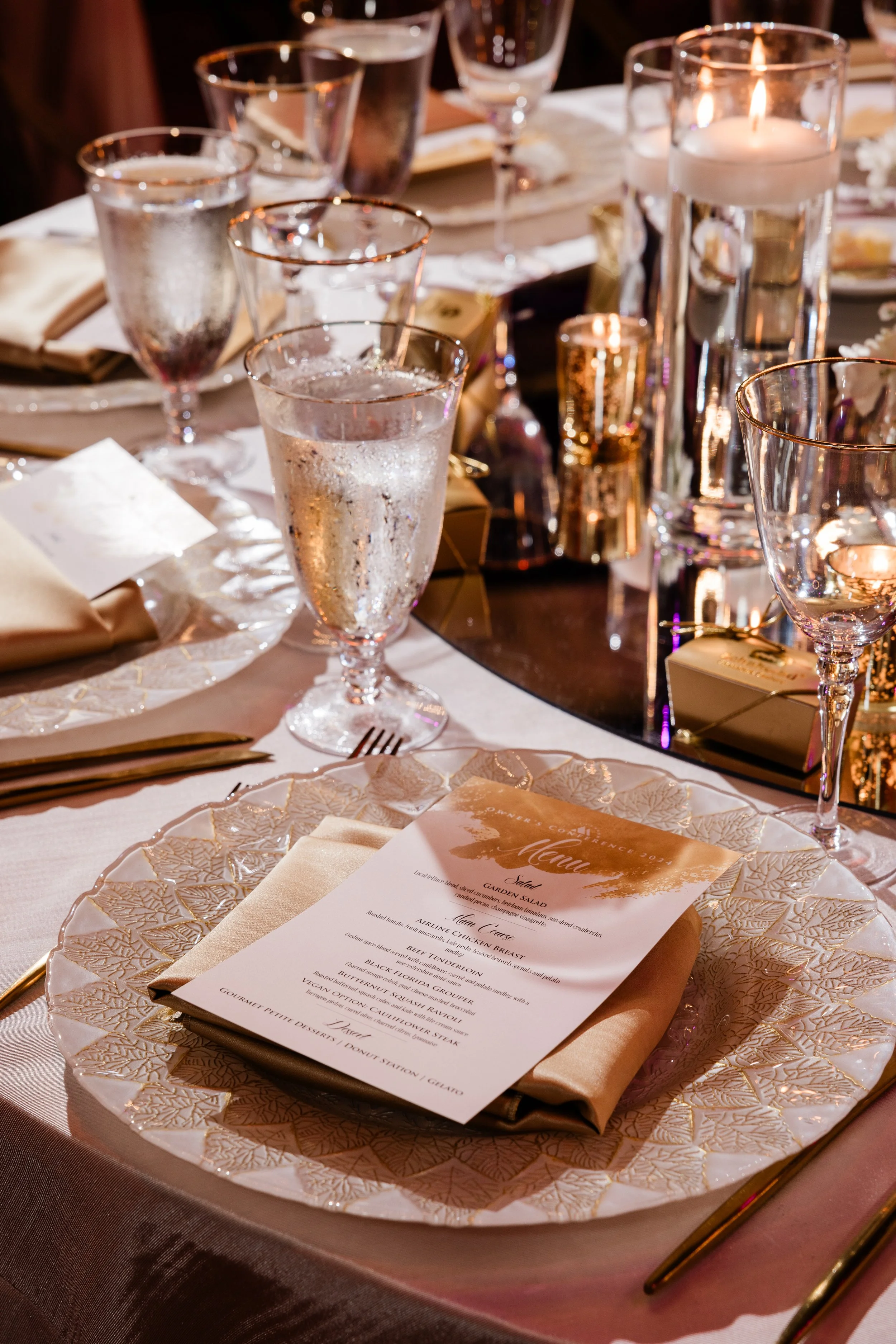 Elegant dining table setting with gold-rimmed glasses of water, gold cutlery, a menu on a decorative plate, and lit candles.
