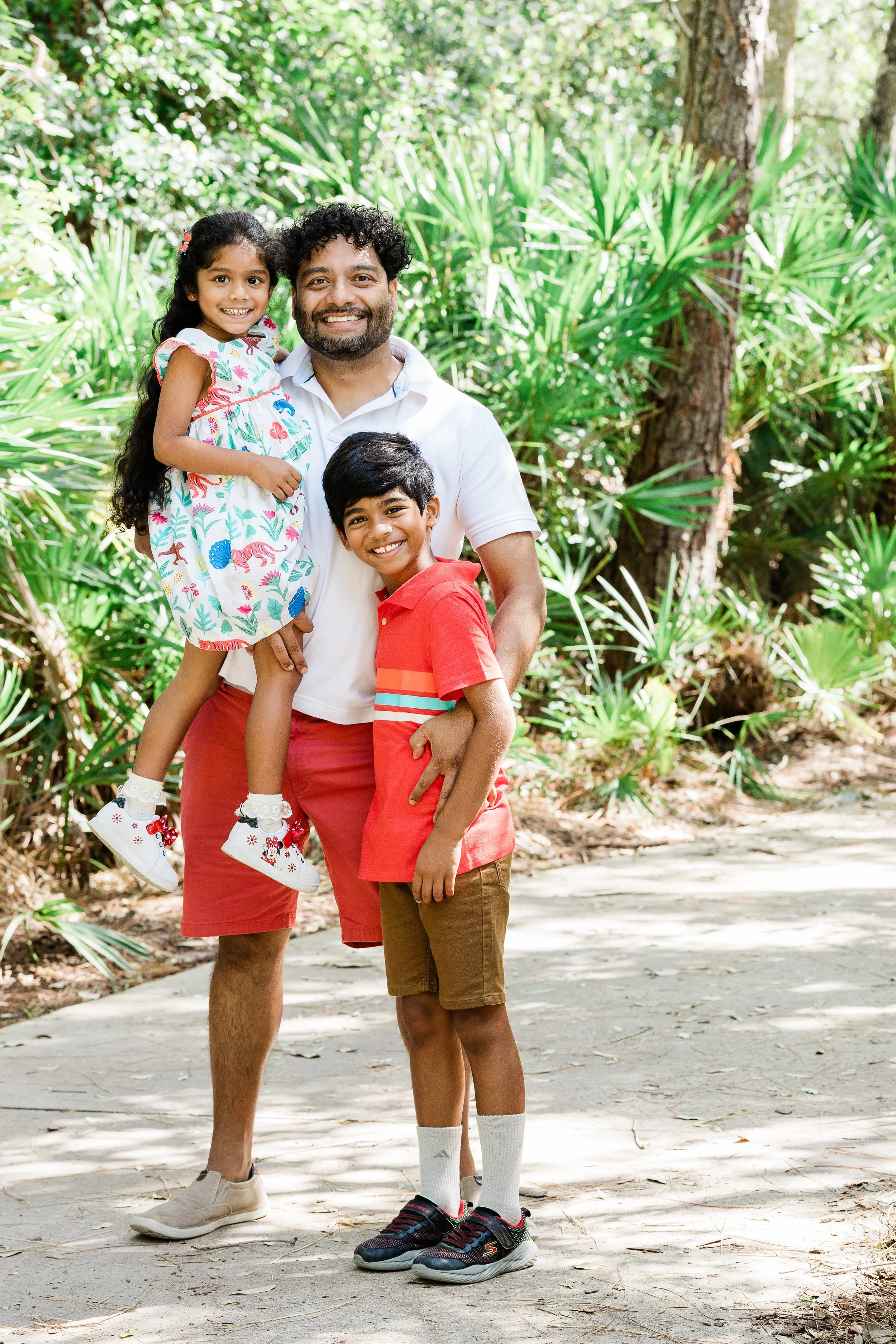 A man and two children standing outdoors on a path surrounded by green plants. The girl is being held by the man, and she is wearing a colorful dress and white sneakers. The boy is standing next to the man, wearing a red shirt, brown shorts, and snea