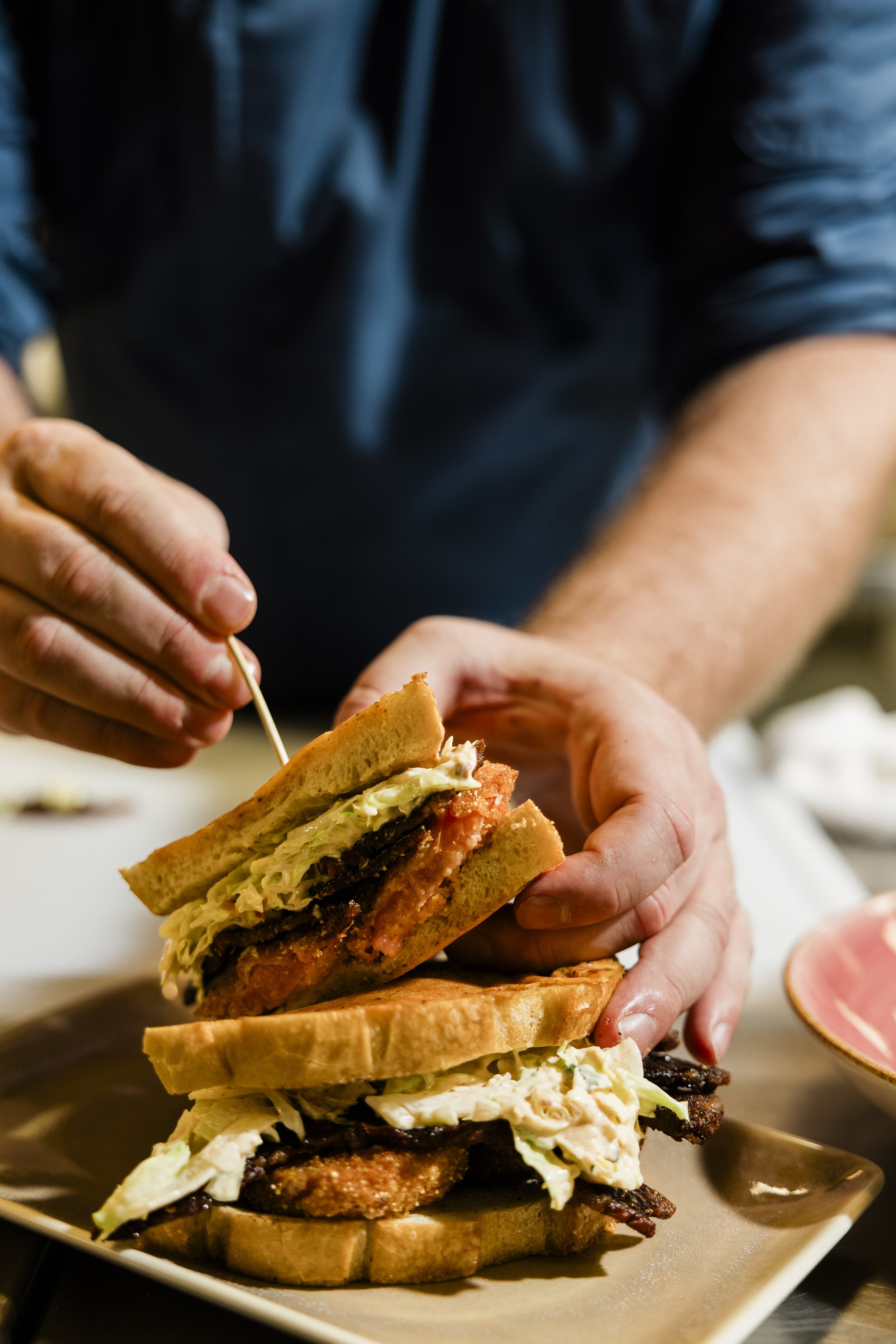 A person prepares to eat a large, multi-layered burger with lettuce, tomato, cheese, and bacon on toasted bread, topped with fried chicken, in a restaurant setting.