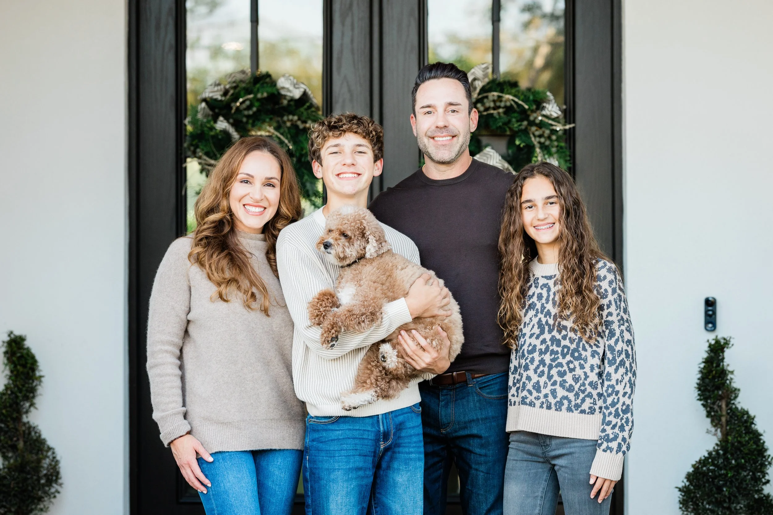 Family of four standing in front of a door decorated with holiday wreaths, with a dog in the son's arms.