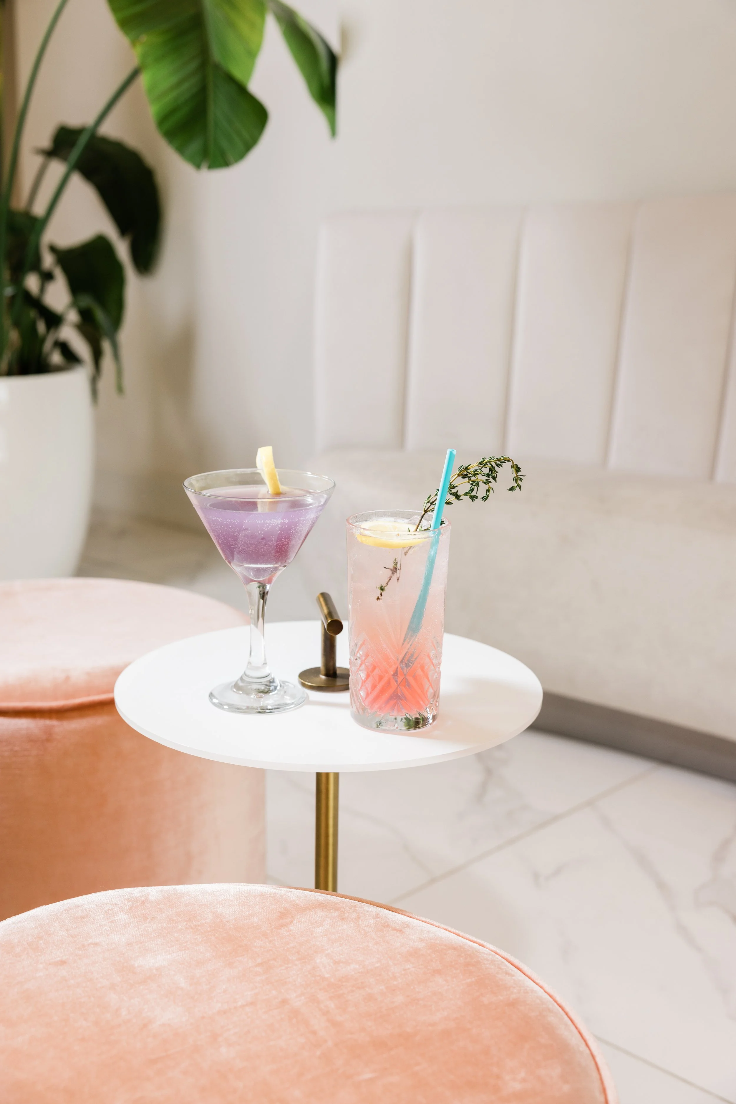 A small white round table with two colorful cocktails, one purple and one pink, each garnished with lemon slices and paper straws. A large green potted plant is in the background, and the setting appears to be indoors with a minimalistic, modern deco