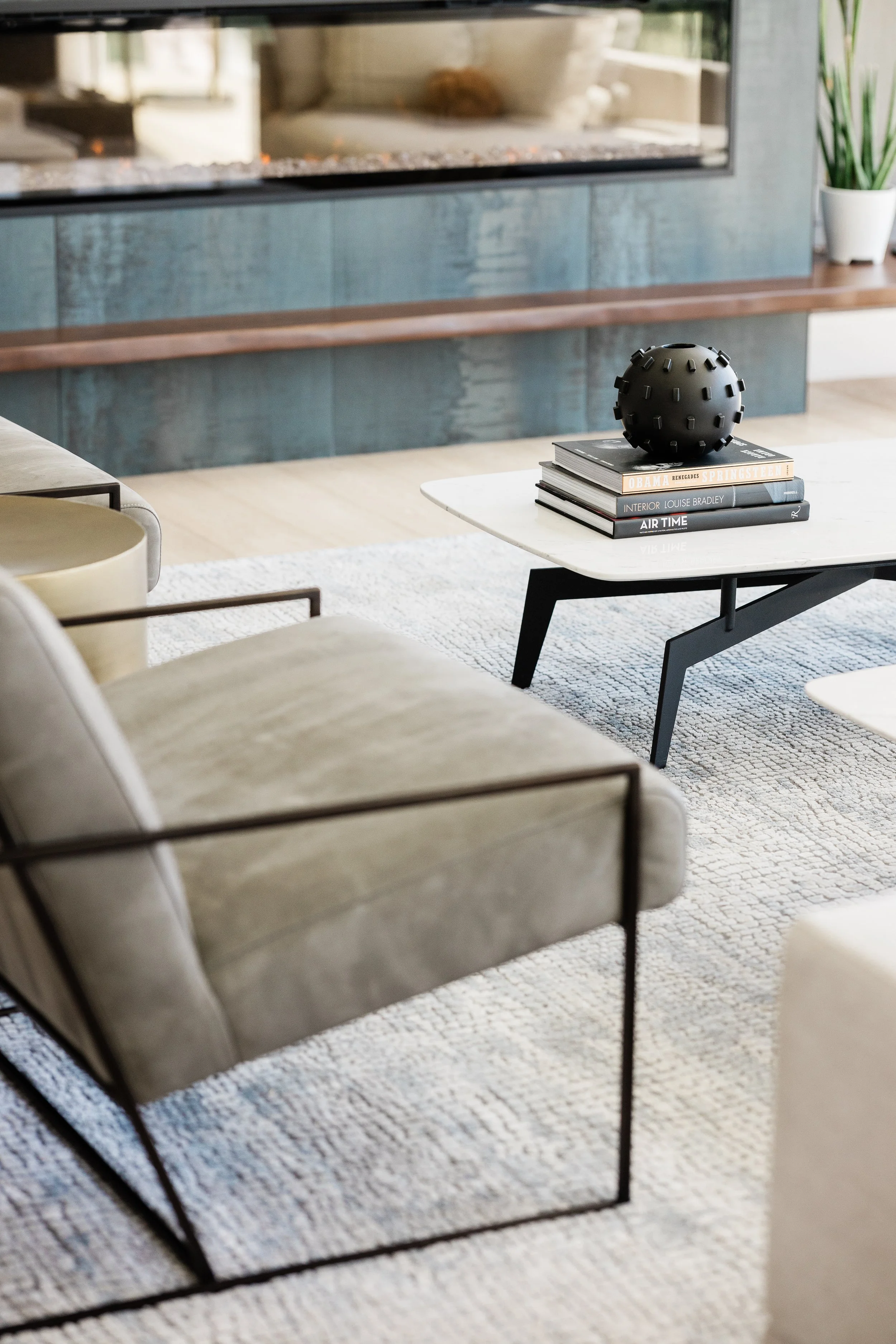 A modern living room with a beige armchair, a white marble coffee table, and a black decorative orb on a stack of books, with a fireplace and a potted plant in the background. Interior design by jamie finley & co.