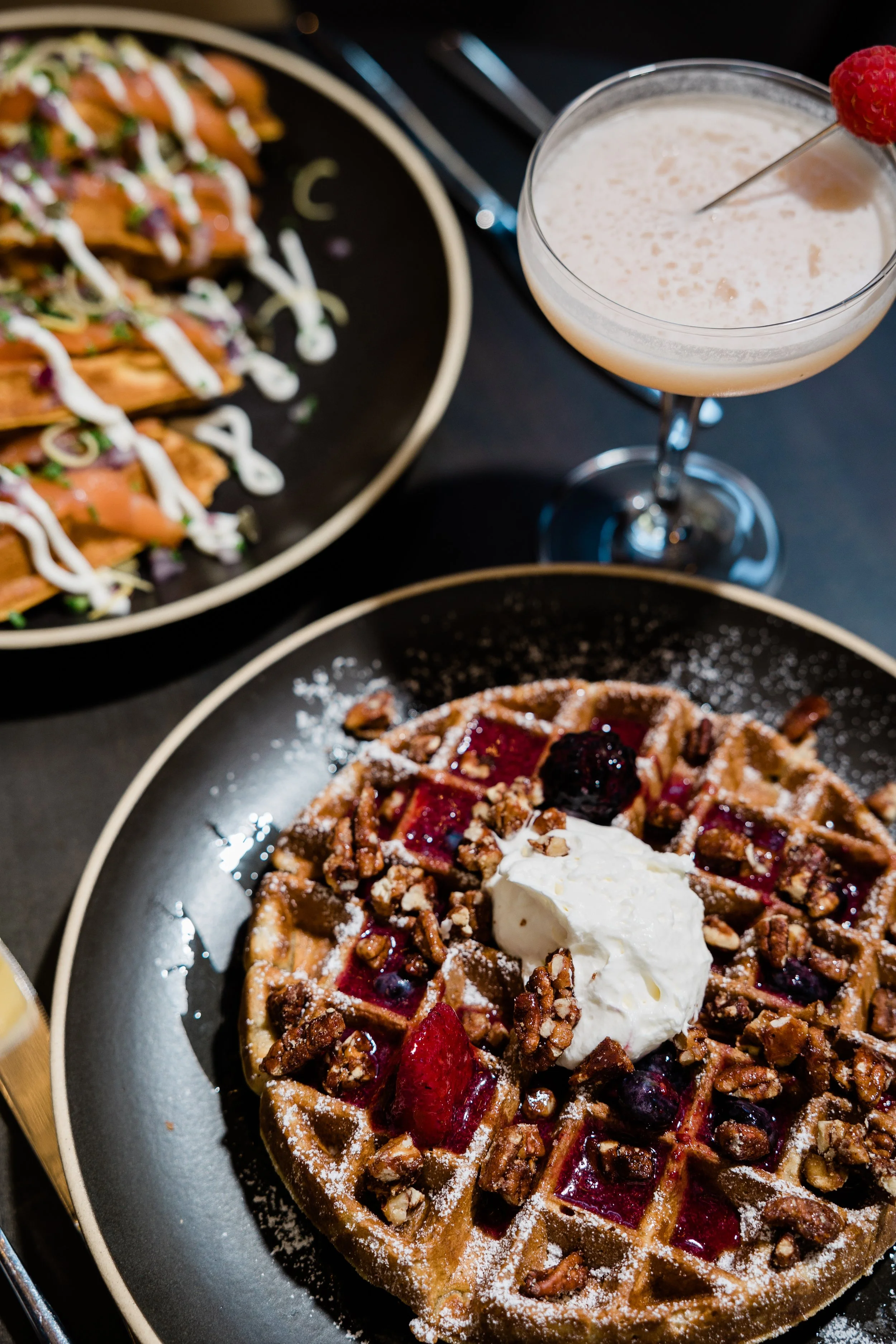 A waffle topped with whipped cream, berries, and chopped nuts, served on a black plate, with a raspberry garnish on a cocktail glass nearby and a plate of salmon tacos with drizzle and vegetables in the background.