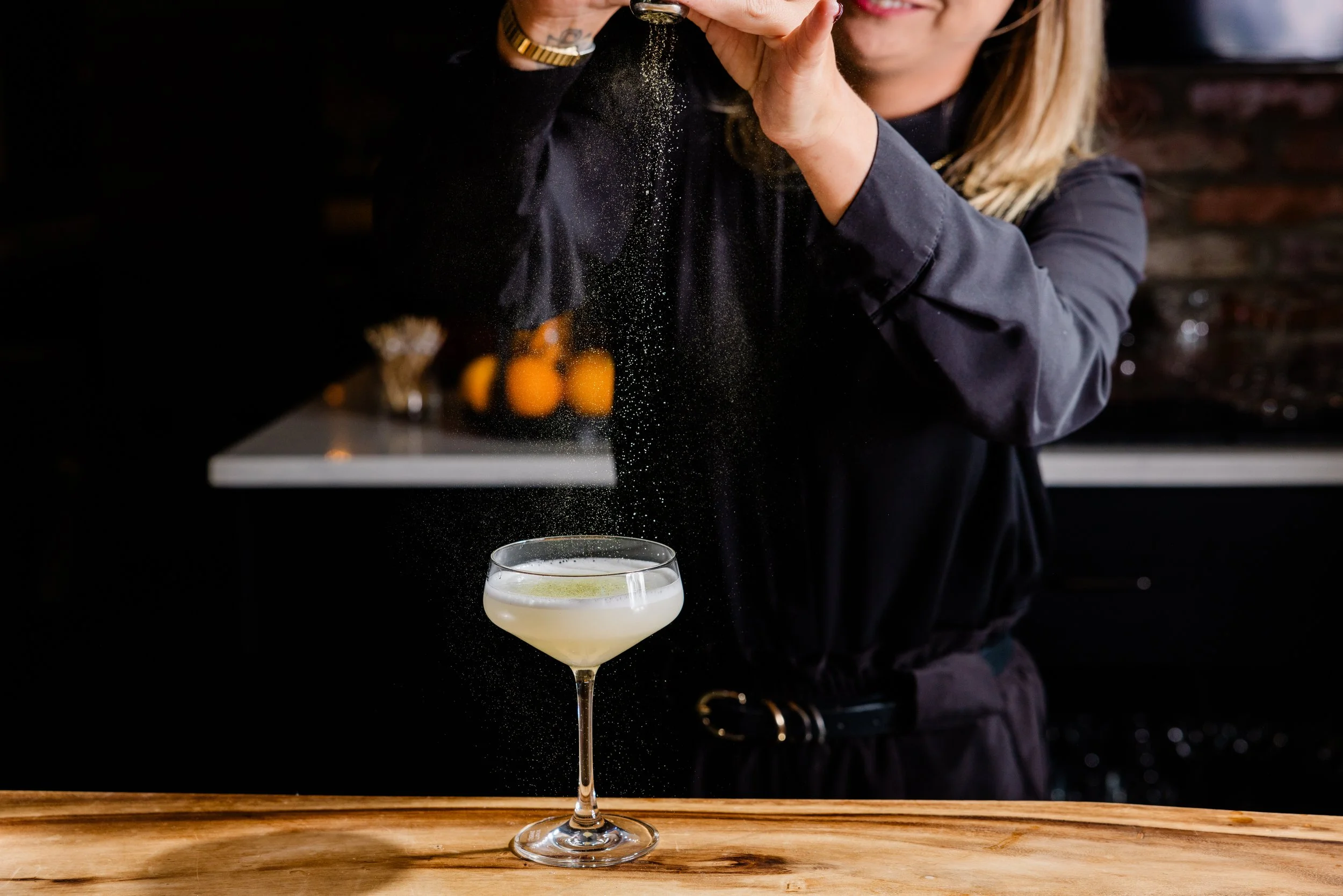 A woman with blonde hair pouring gold-colored powder from a shaker into a cocktail glass that is filled with a pale yellow liquid and garnished with a lime wedge. The scene appears to be in a bar or restaurant setting with a dark background.