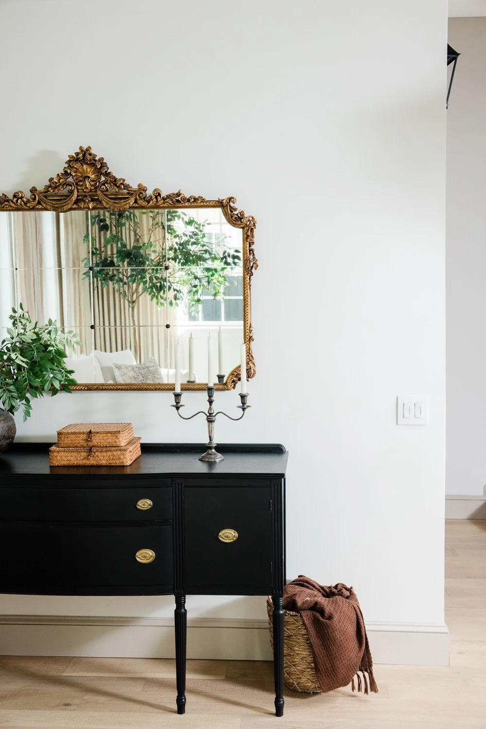 A black dresser with gold handles, decorated with a silver candelabra with white candles, a potted green plant, and two woven boxes. A large, ornate gold-framed mirror hangs above it. A wicker basket with a brown blanket is on the floor beside the dr