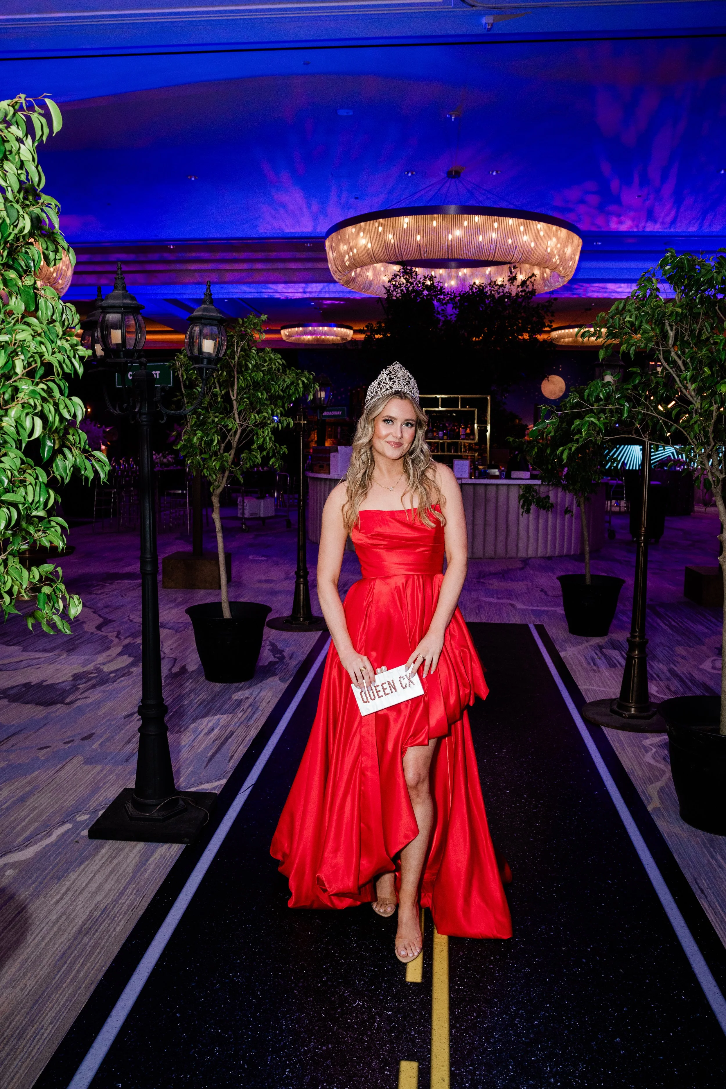 A woman in a red dress and tiara standing on a black aisle with a yellow line in a decorated interior with plants, vintage lamps, and ornate chandeliers, holding a sign that reads 'QUEEN CK'.