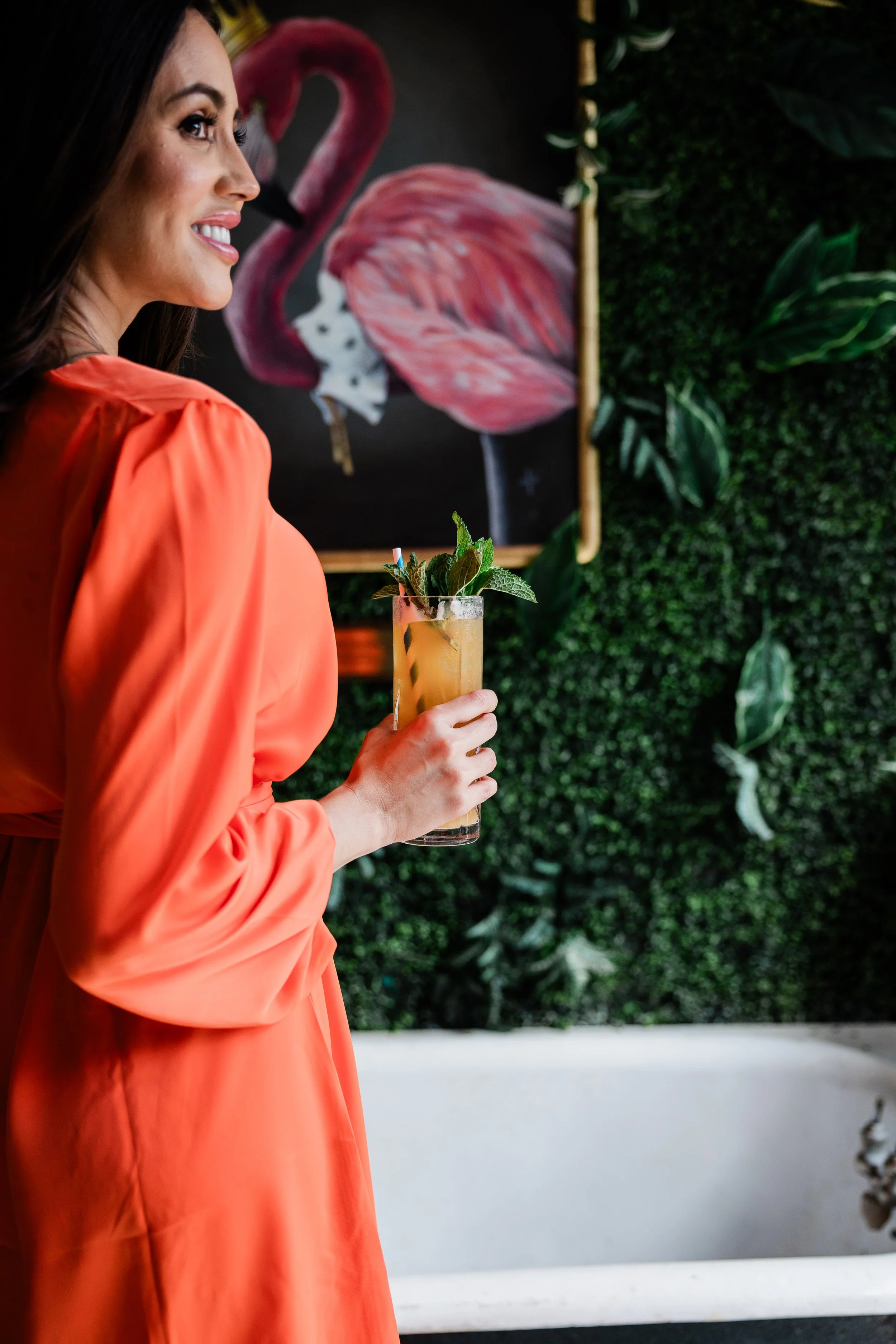 A woman in an orange dress holding a cocktail with mint leaves, standing in front of a wall with a pink flamingo painting and green foliage.