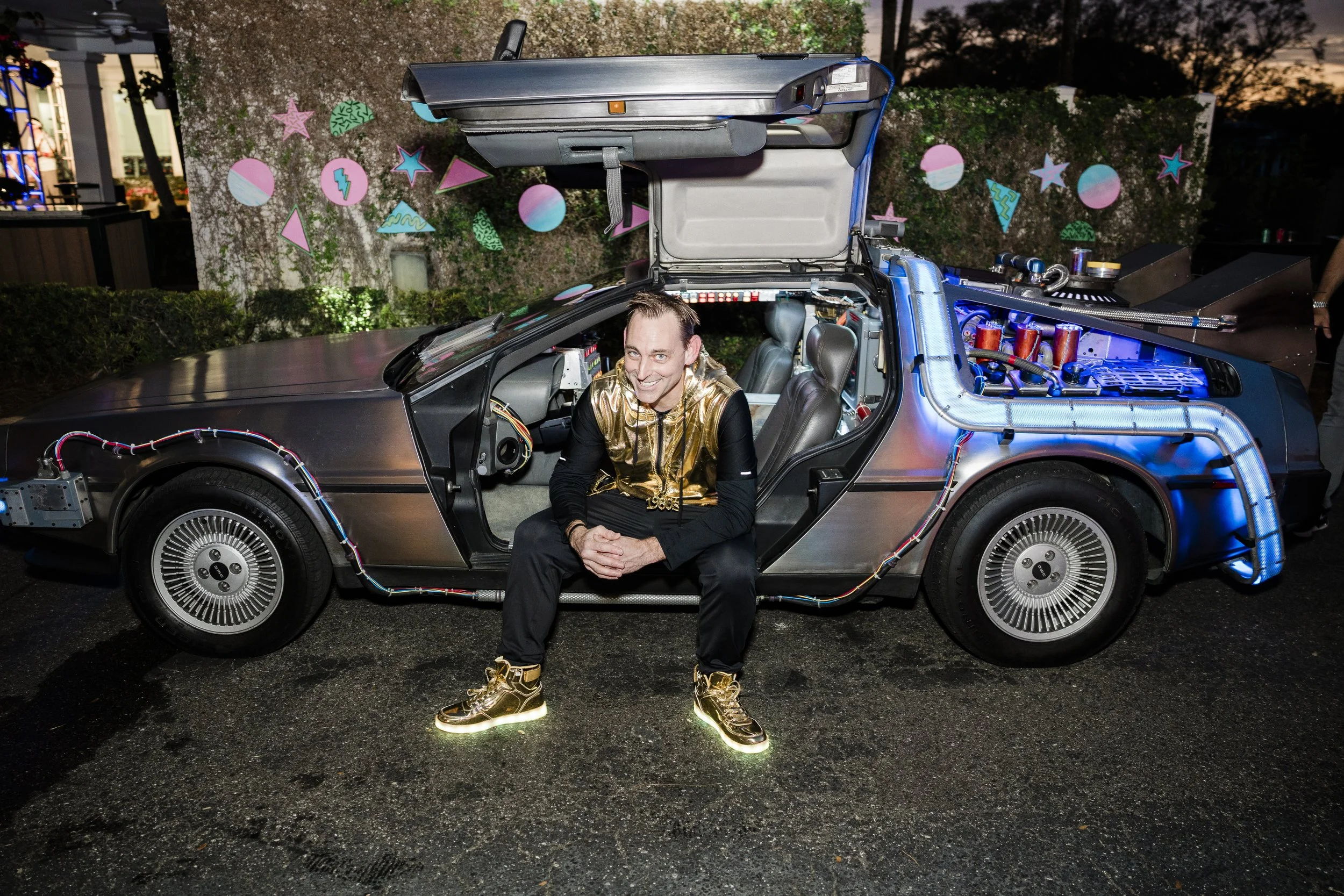 A man in gold jacket and gold sneakers sitting on the ground in front of a DeLorean car with its door open, exposing the interior and some electronic components. The scene is from the Heart Gallery fundraiser at Palma Ceia Golf Club.