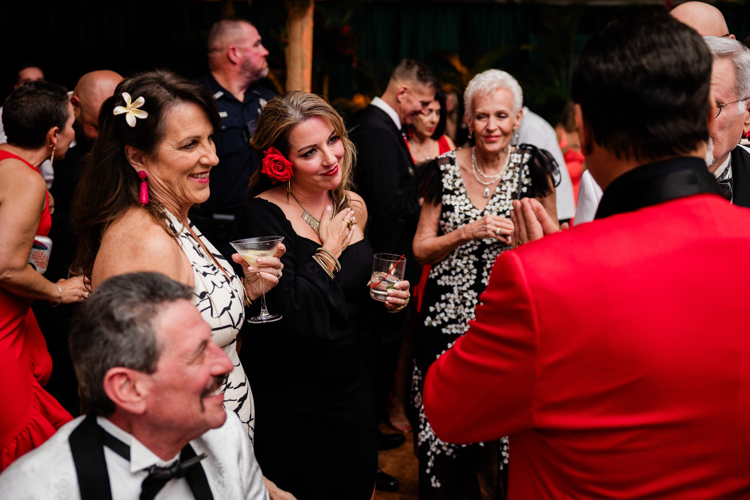 People dressed in formal attire, some with flowers in their hair, socializing and smiling at a festive event. One woman is holding a cocktail glass, and others are engaged in conversation, indicating a lively gathering or celebration.