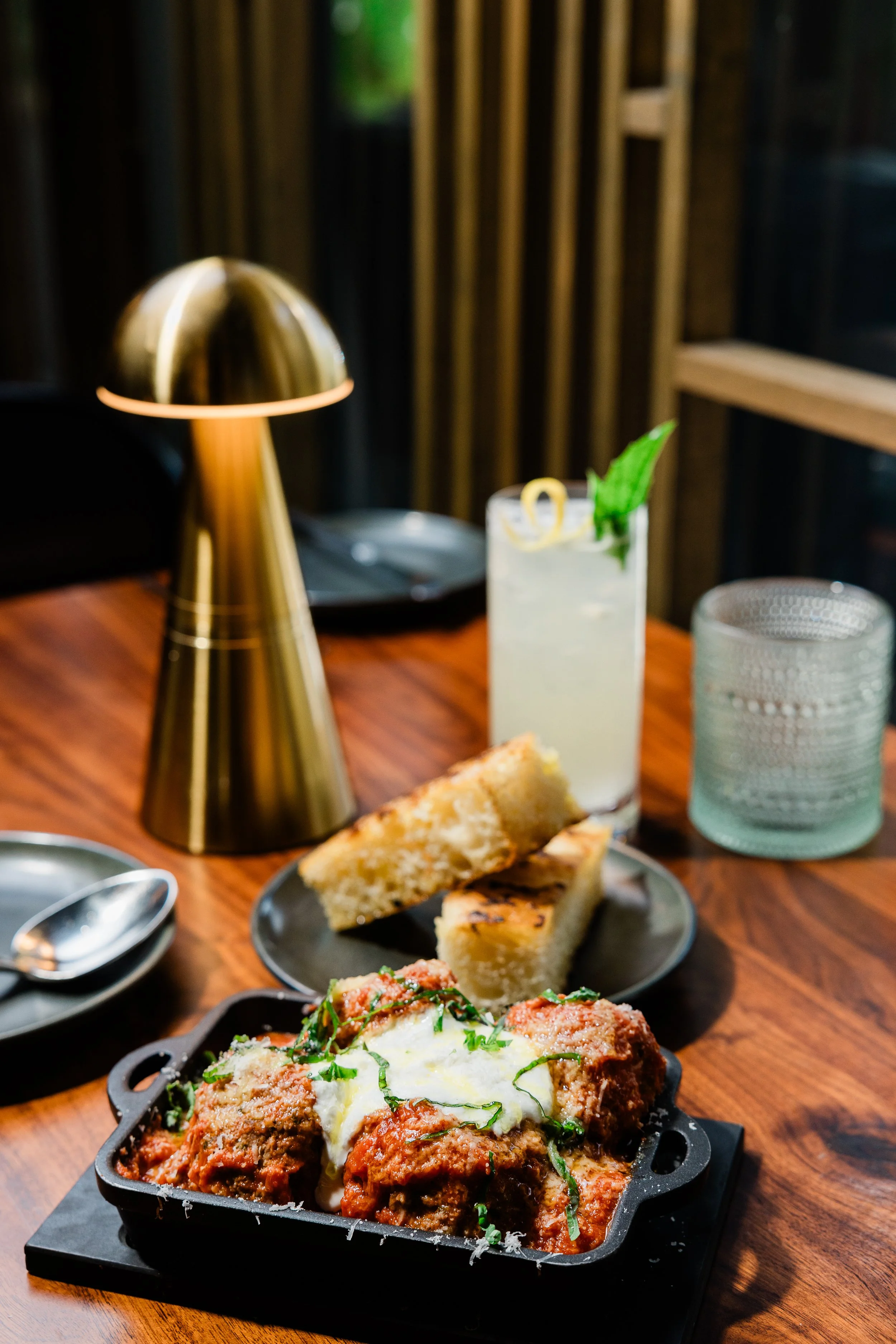 A table with a dish of baked Italian meatball parmesan topped with melted cheese and garnished with herbs, slices of garlic bread, and a drink with lemon slice and mint, with a gold lamp and a wooden wall in the background.