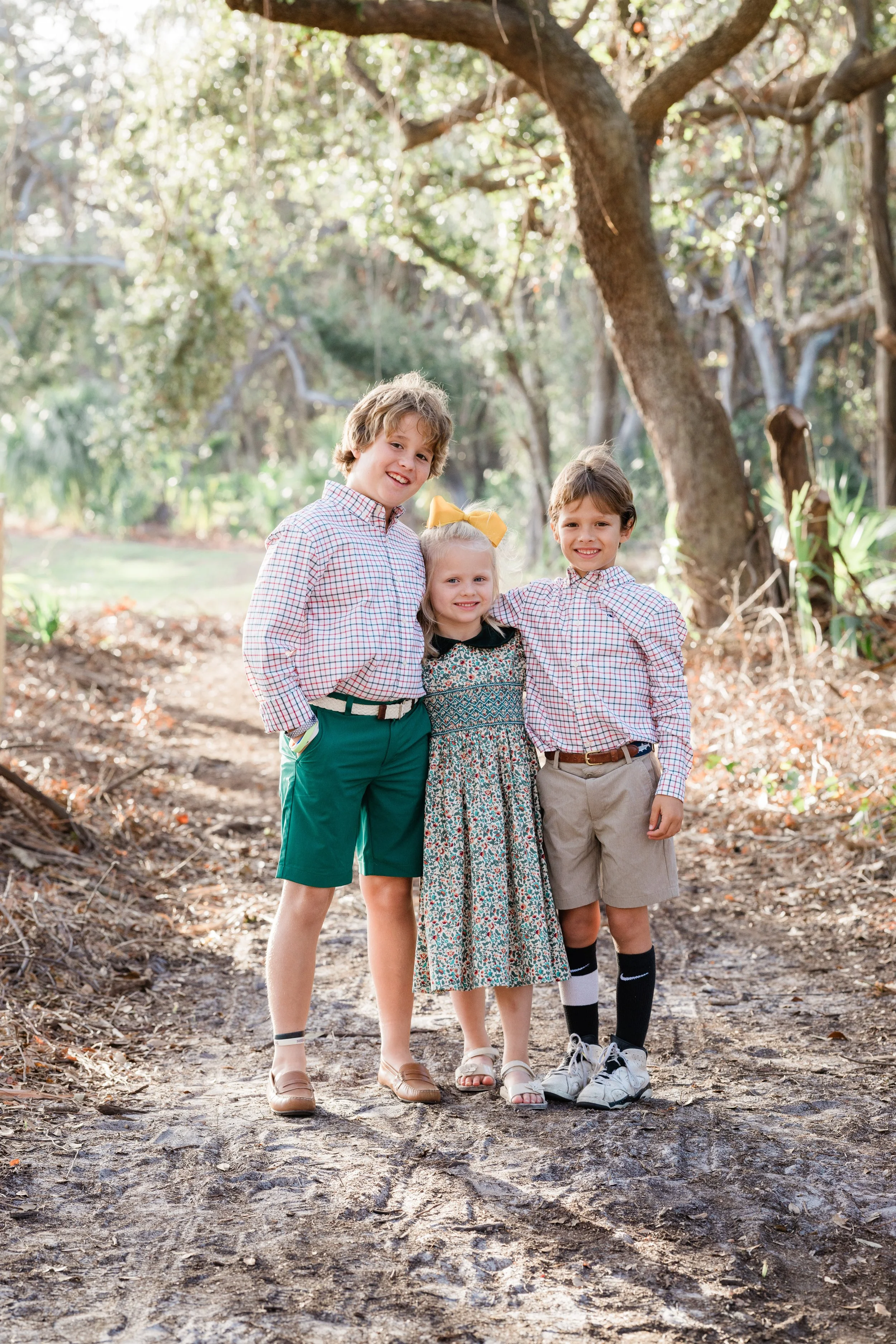 Four children standing side by side outdoors on a dirt path with trees and sunlight in the background. They are smiling and dressed in casual clothing, with two boys and two girls.