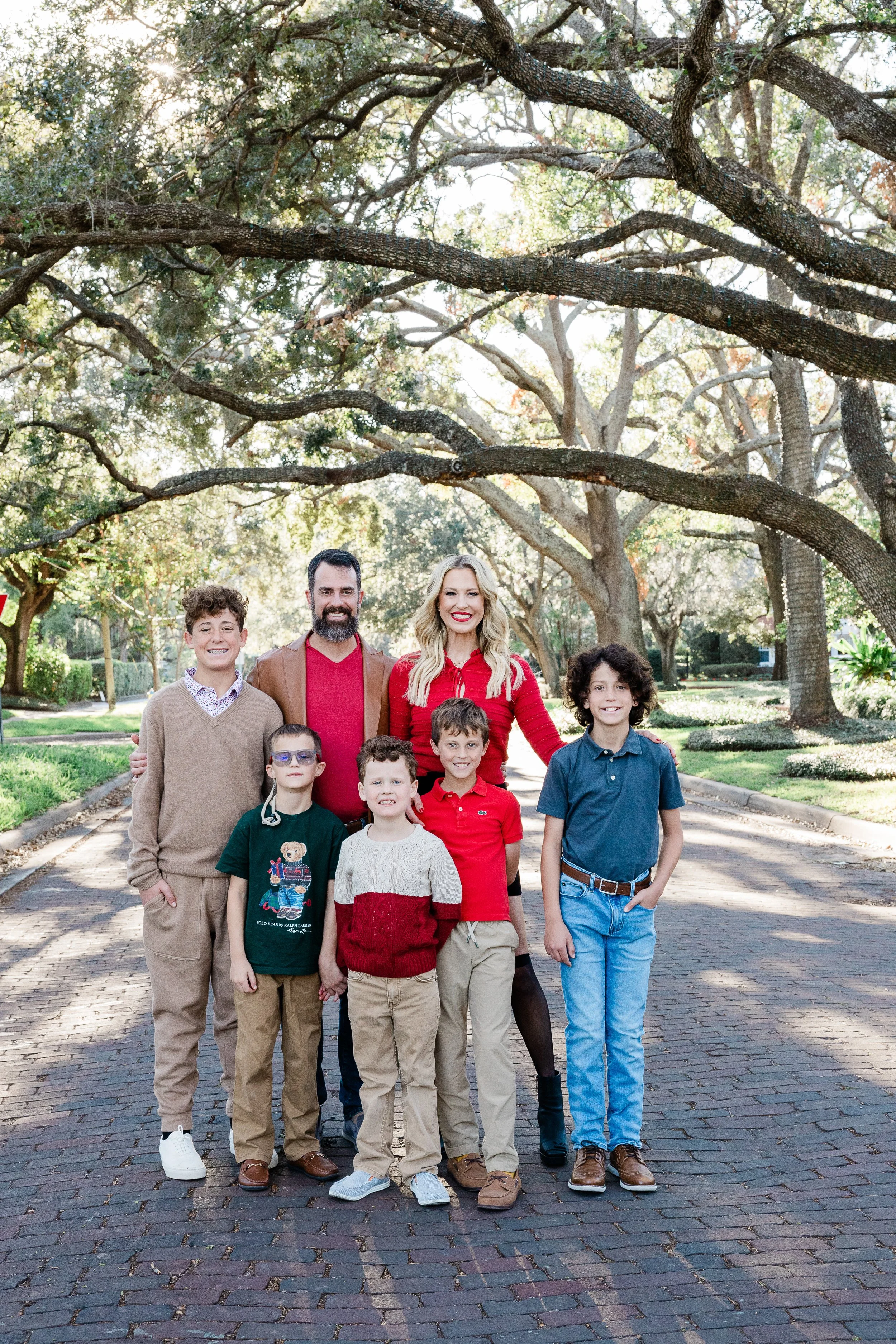 A group of seven people, including four children and three adults, standing outdoors on a brick street surrounded by large trees with broad branches and green foliage, smiling and posing for the photo.