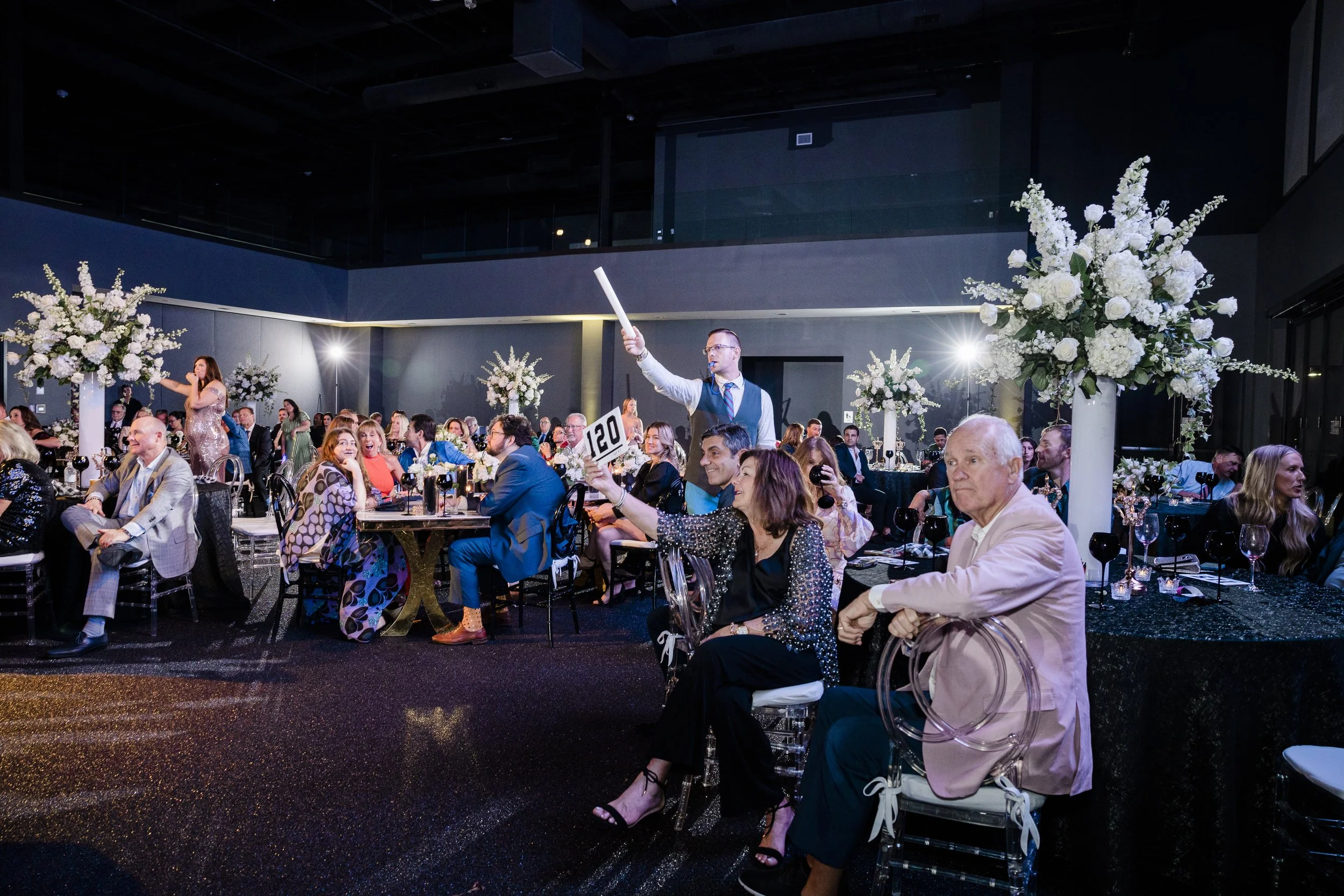 A large group of people seated at round tables decorated with floral centerpieces at a formal event or celebration in a modern venue.