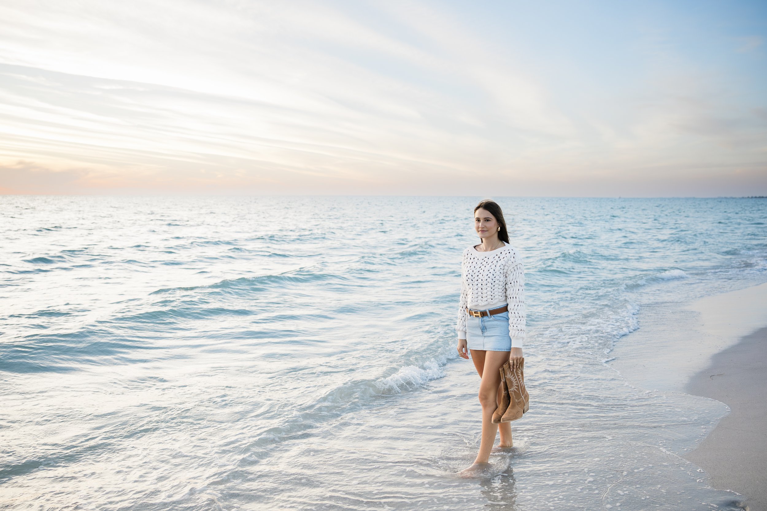 Young woman standing waist-deep in the ocean at sunset, wearing a white knit sweater and light blue shorts, holding brown boots in her hand.