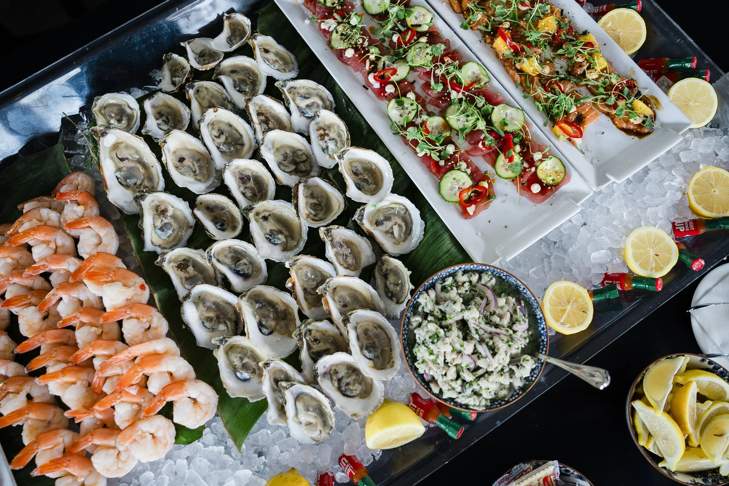 Assorted seafood platter with oysters on a bed of ice, shrimp, and colorful garnished tuna and salmon sashimi with lemon slices, surrounded by dipping sauces and hot sauce bottles.