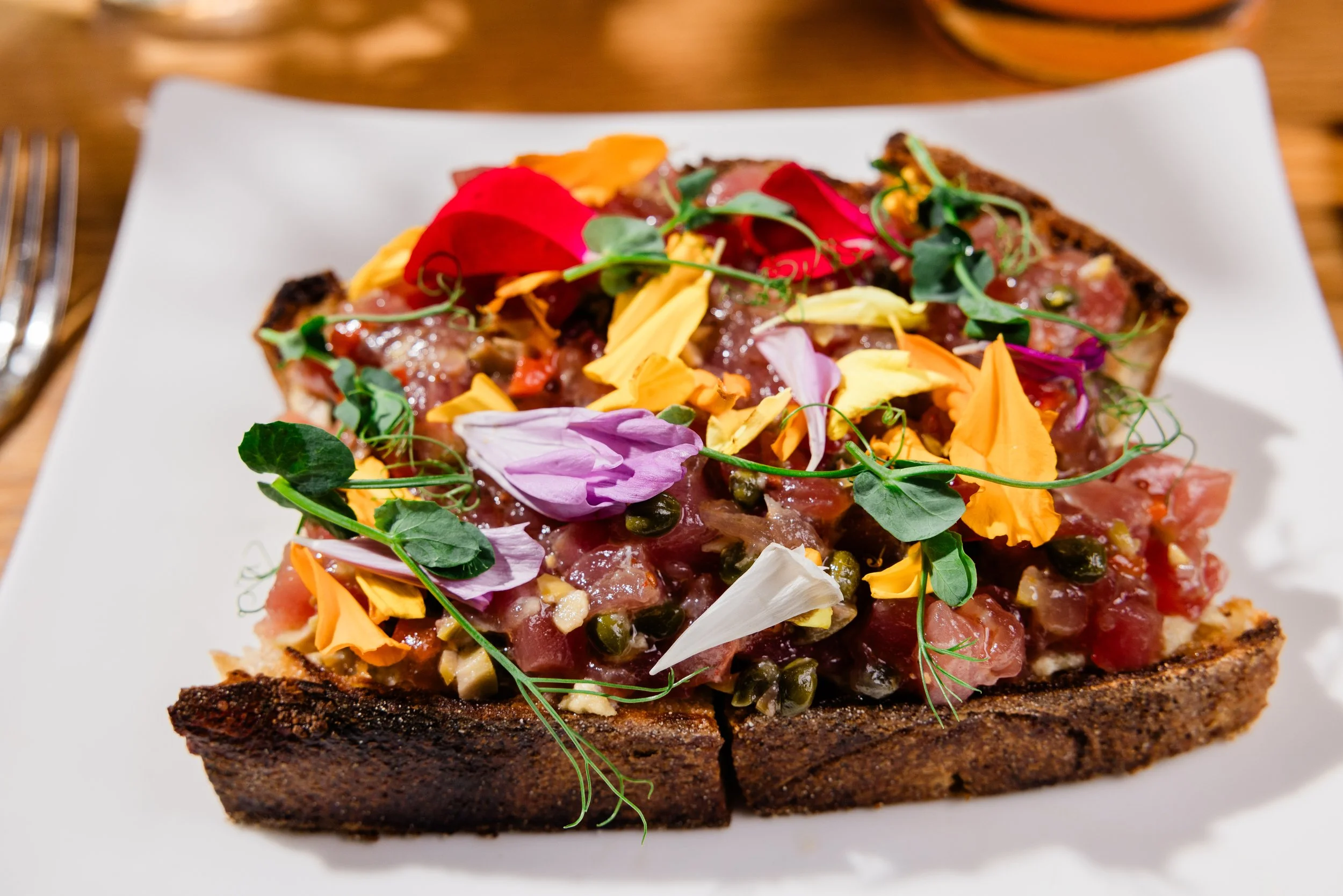 A slice of toasted bread topped with assorted colorful edible flowers, herbs, and green peas on a white plate.