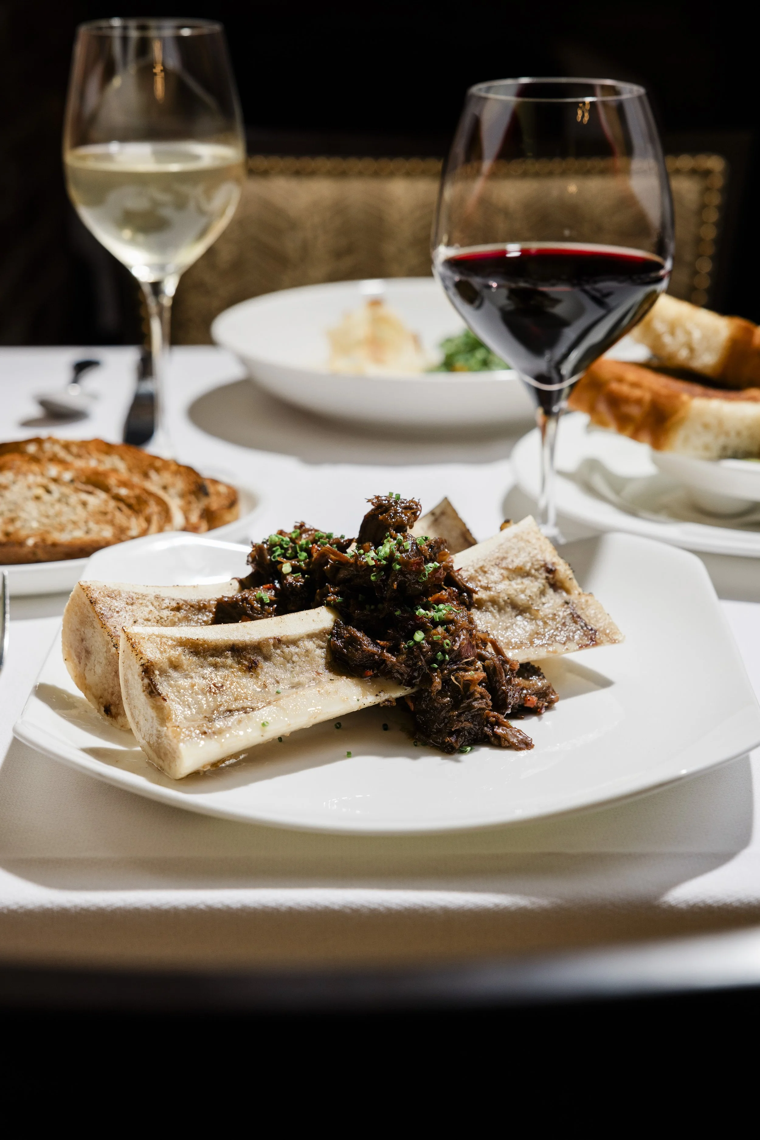 A plate of sliced beef served with a dark sauce and garnished with chopped herbs, with a glass of red wine and a glass of white wine on a table set for dinner.