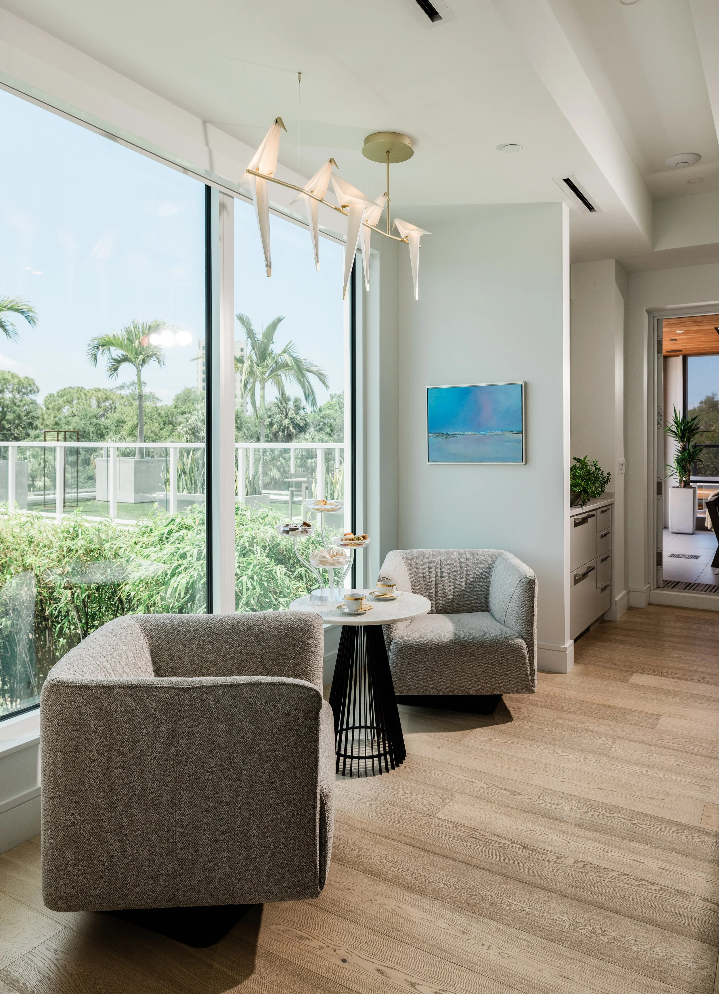 A bright, modern living area with large floor-to-ceiling windows showing palm trees and greenery outside. The room features a small round table with teacups, a light-colored sofa, and a piece of wall art depicting a serene landscape. Ceiling lights a