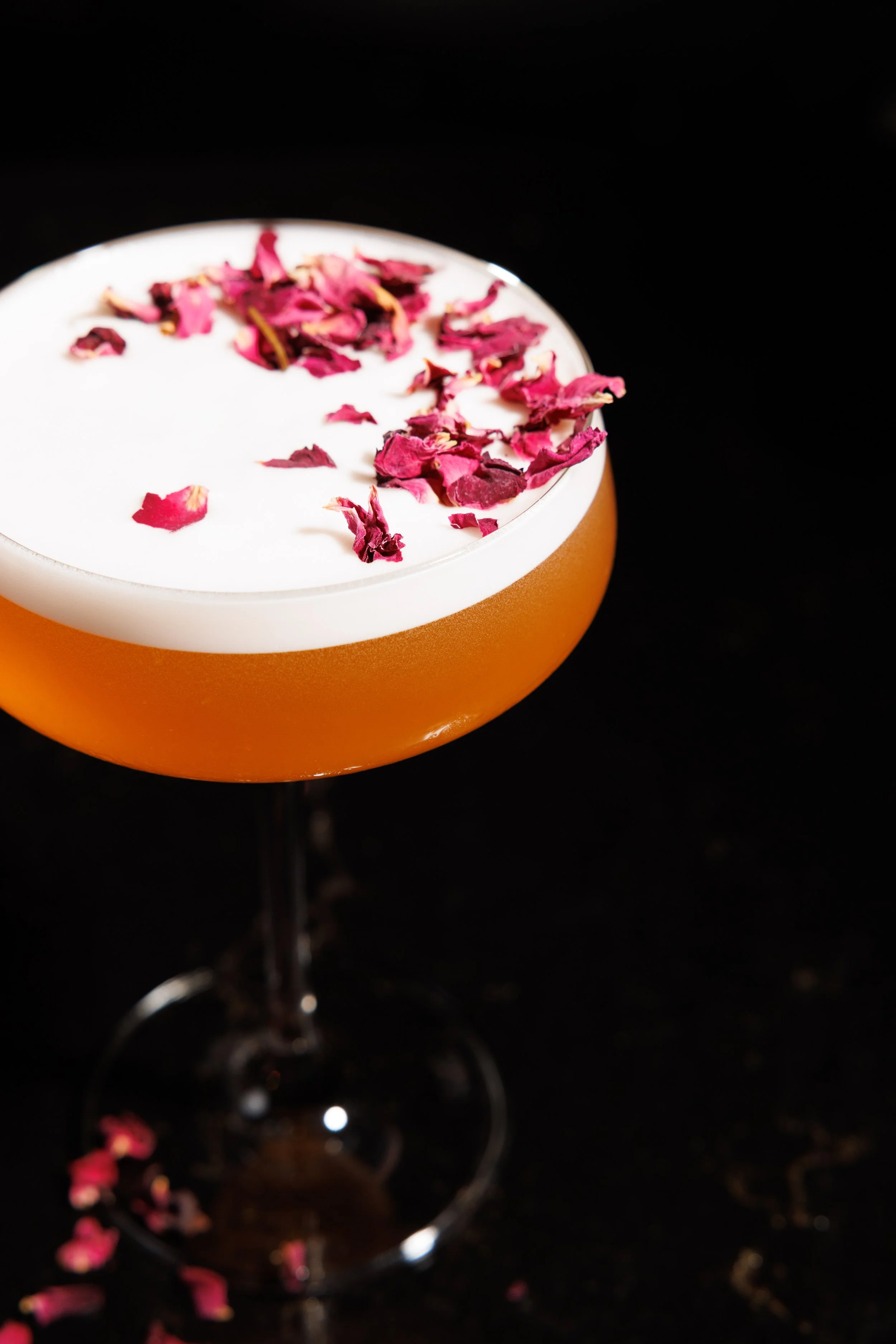 Cocktail glass with orange-colored drink topped with white foam and pink rose petals, against a black background.