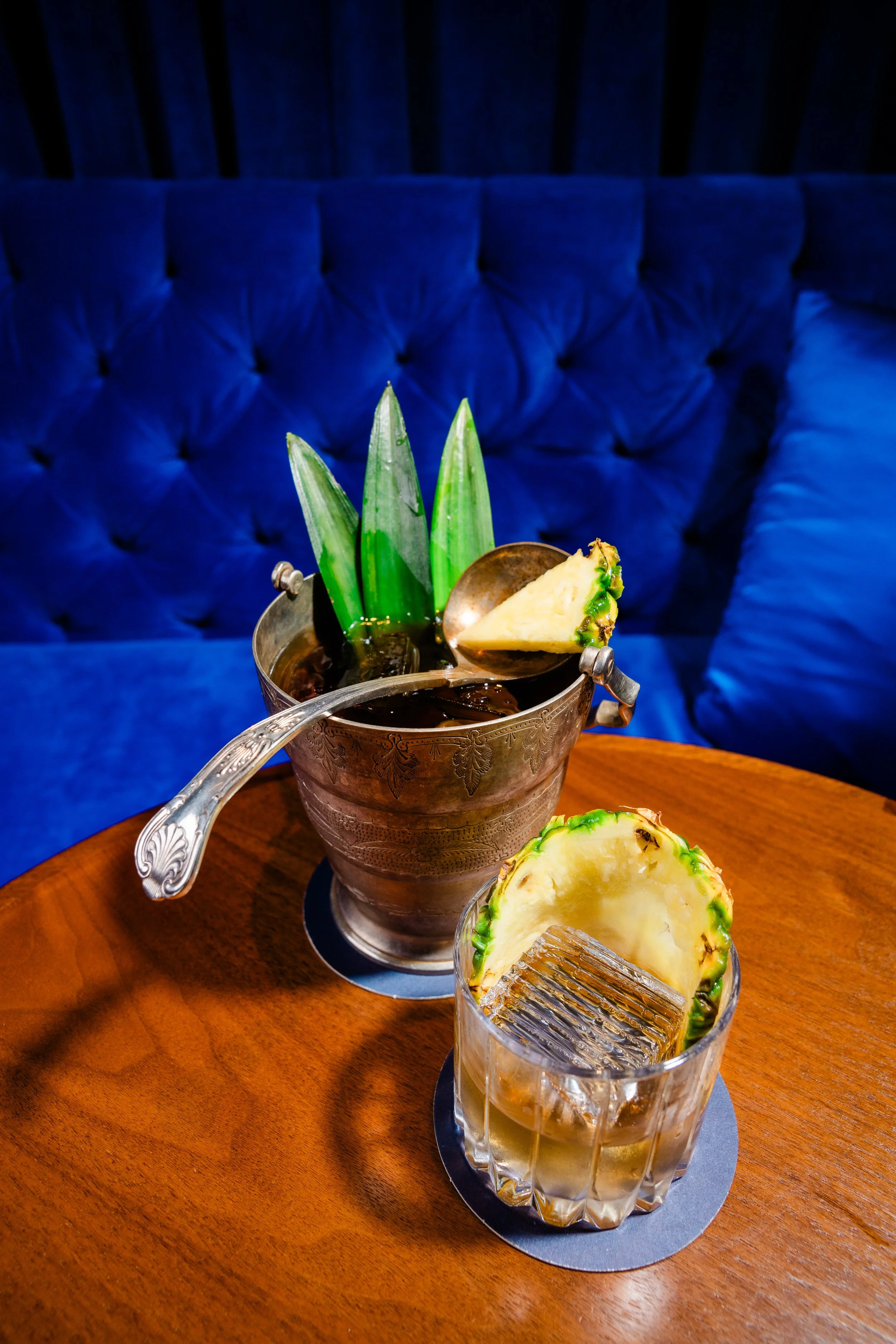 A pewter cocktail pitcher garnished with pineapple leaves, filled with ice cubes and a cocktail. An antique ladle balances on the rim with a slice of pineapple. Set on a wooden table with a dark blue tufted velvet background. Photographed at The Tamp