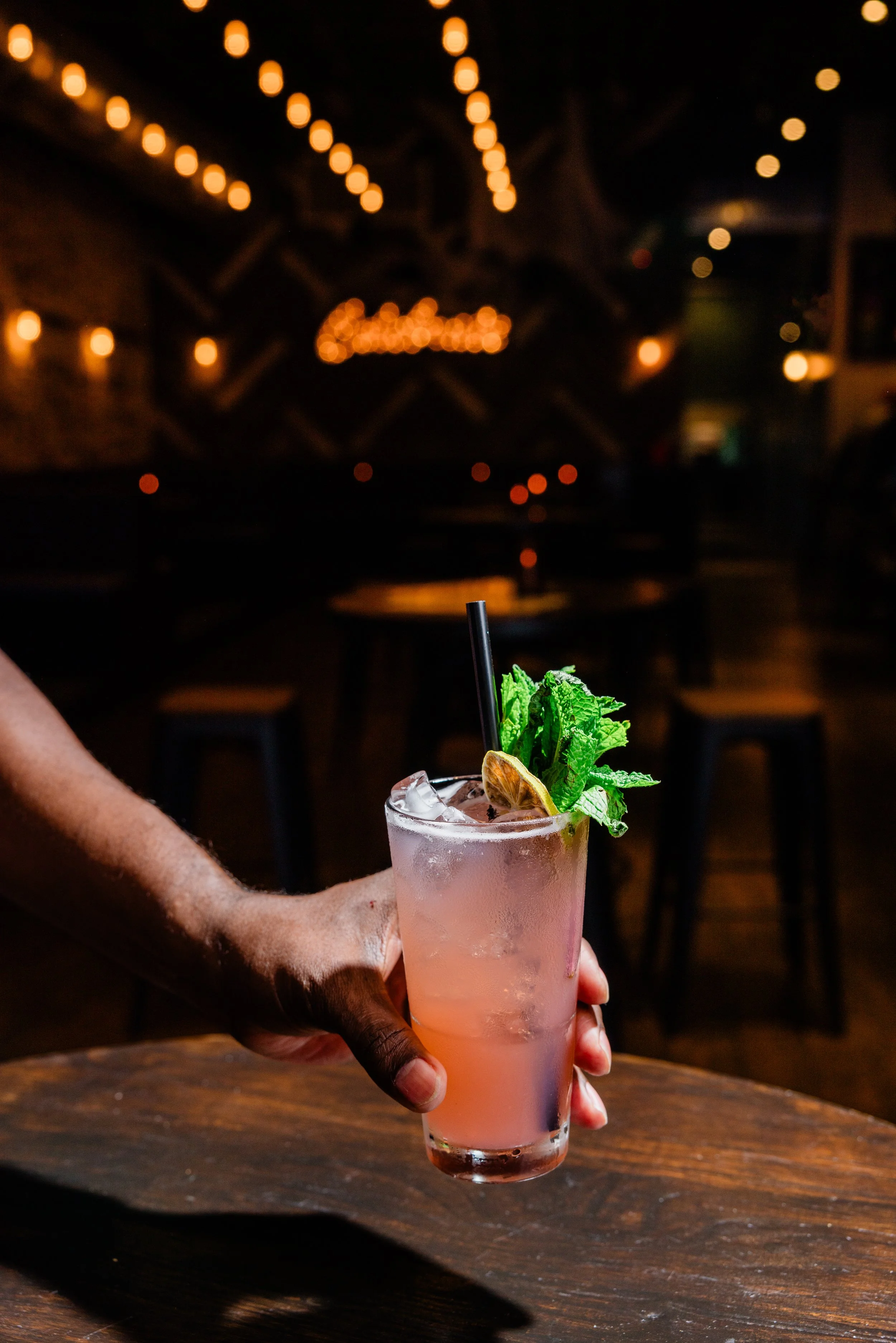 A person holding a pink cocktail garnished with mint leaves and a lemon slice, in a tall glass with ice, in a dimly lit restaurant or bar setting.
