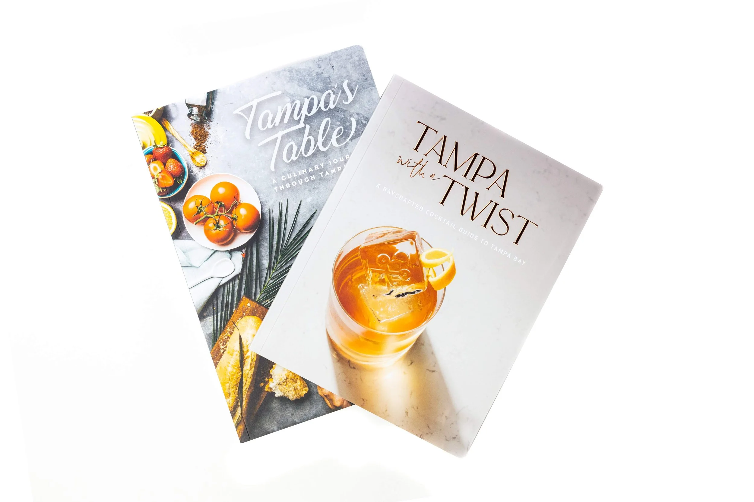 Two cookbooks, one titled 'Tampa's Table' and the other 'Tampa With a Twist,' placed on a white surface.