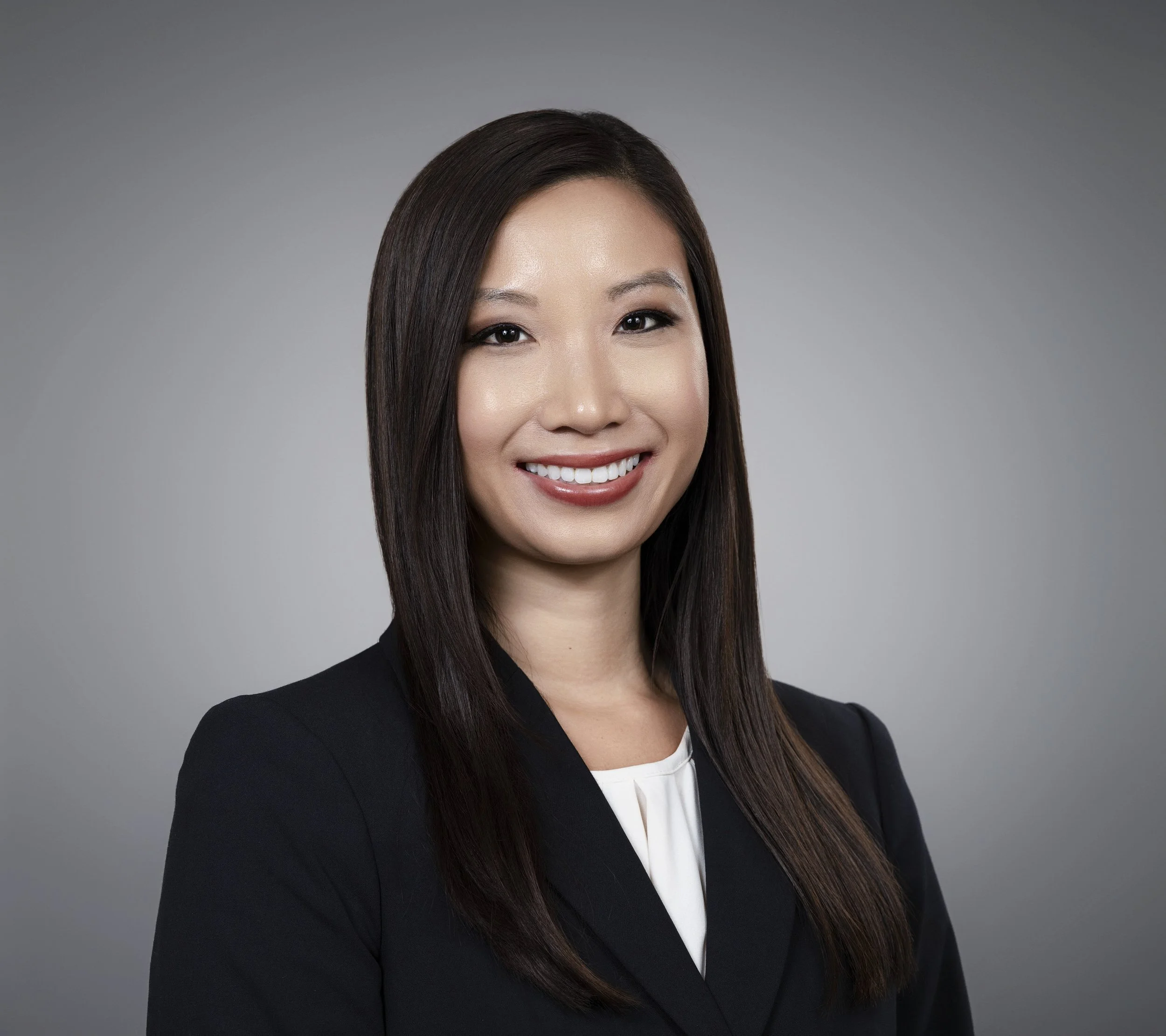 Close-up of a woman with long dark hair, smiling, wearing a black blazer and white top, against a neutral gray background.