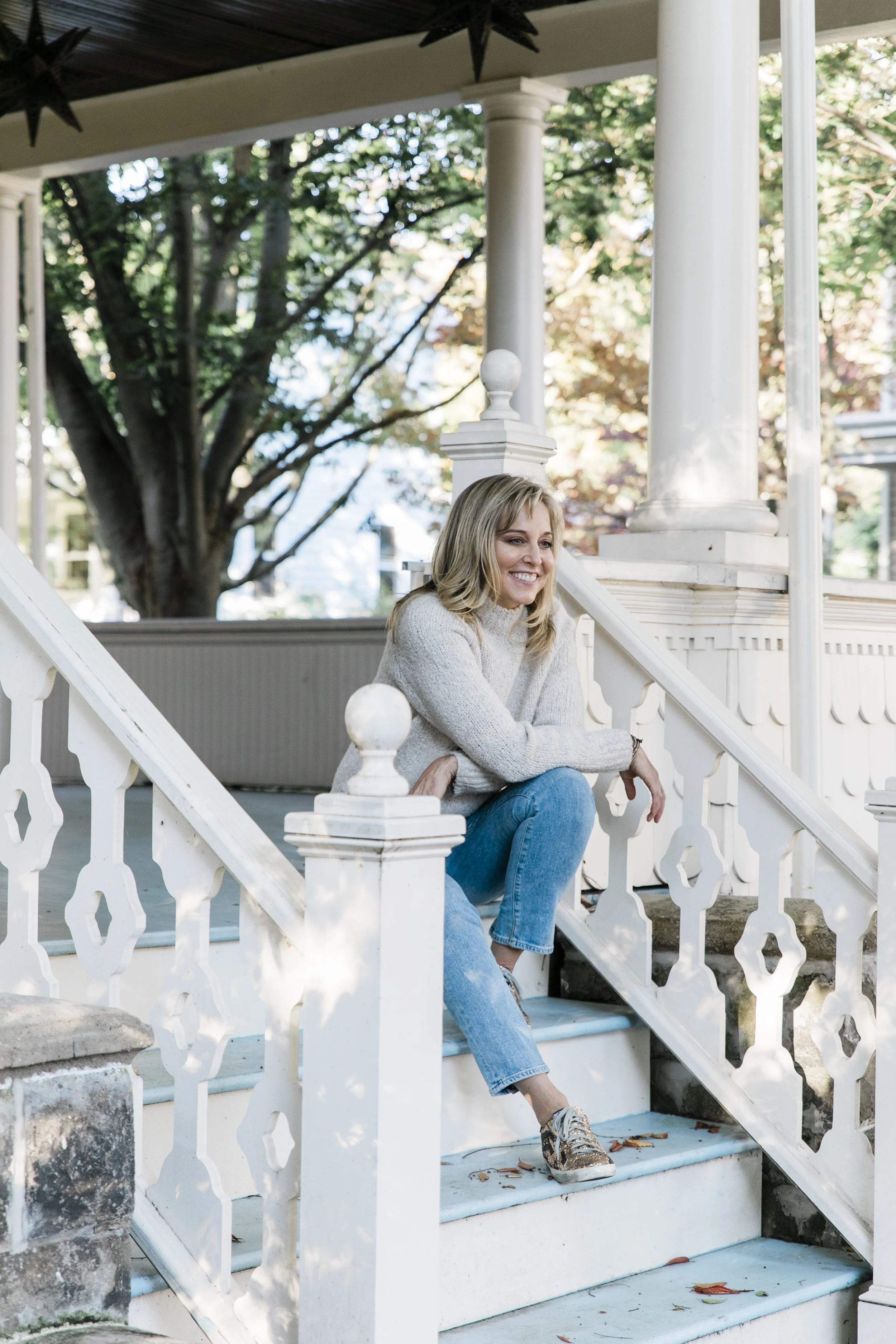 blonde woman in a white sweater and blue jeans sitting on the steps of a porch in autumn in NJ