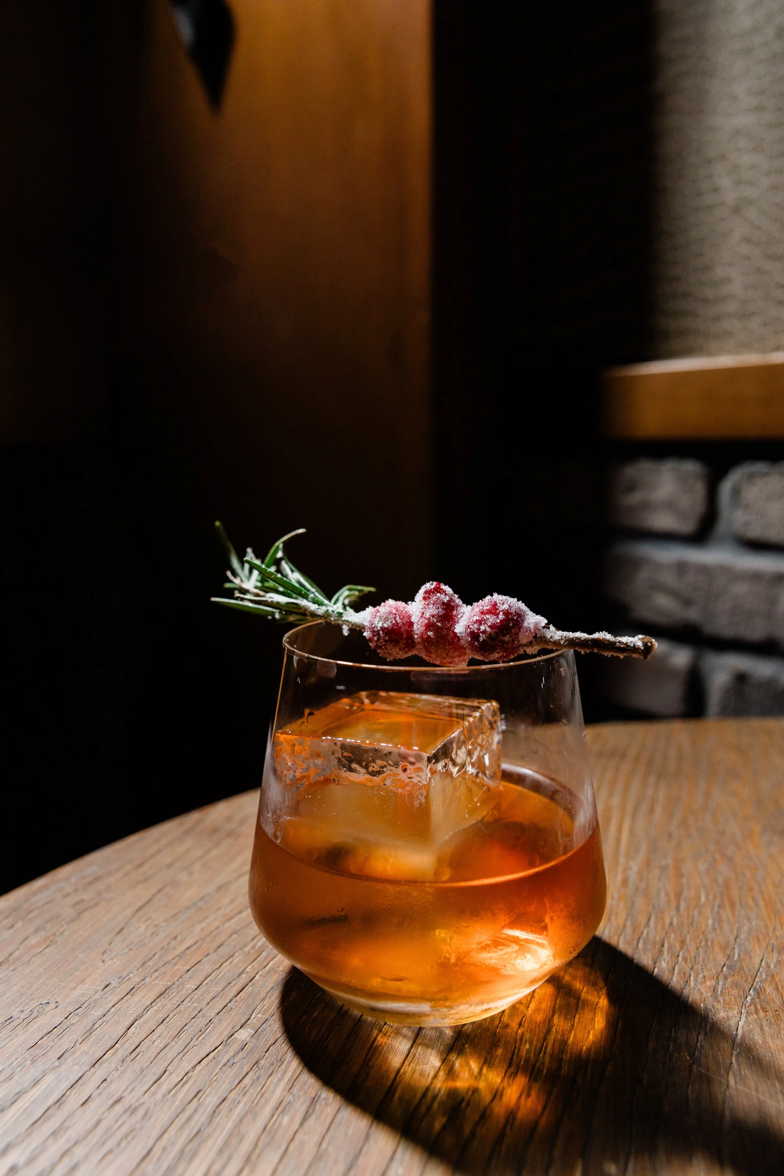 A glass of amber-colored beverage with ice cubes, garnished with a sprig of rosemary and a frosted red berry branch on top, placed on a wooden table in a dimly lit setting.