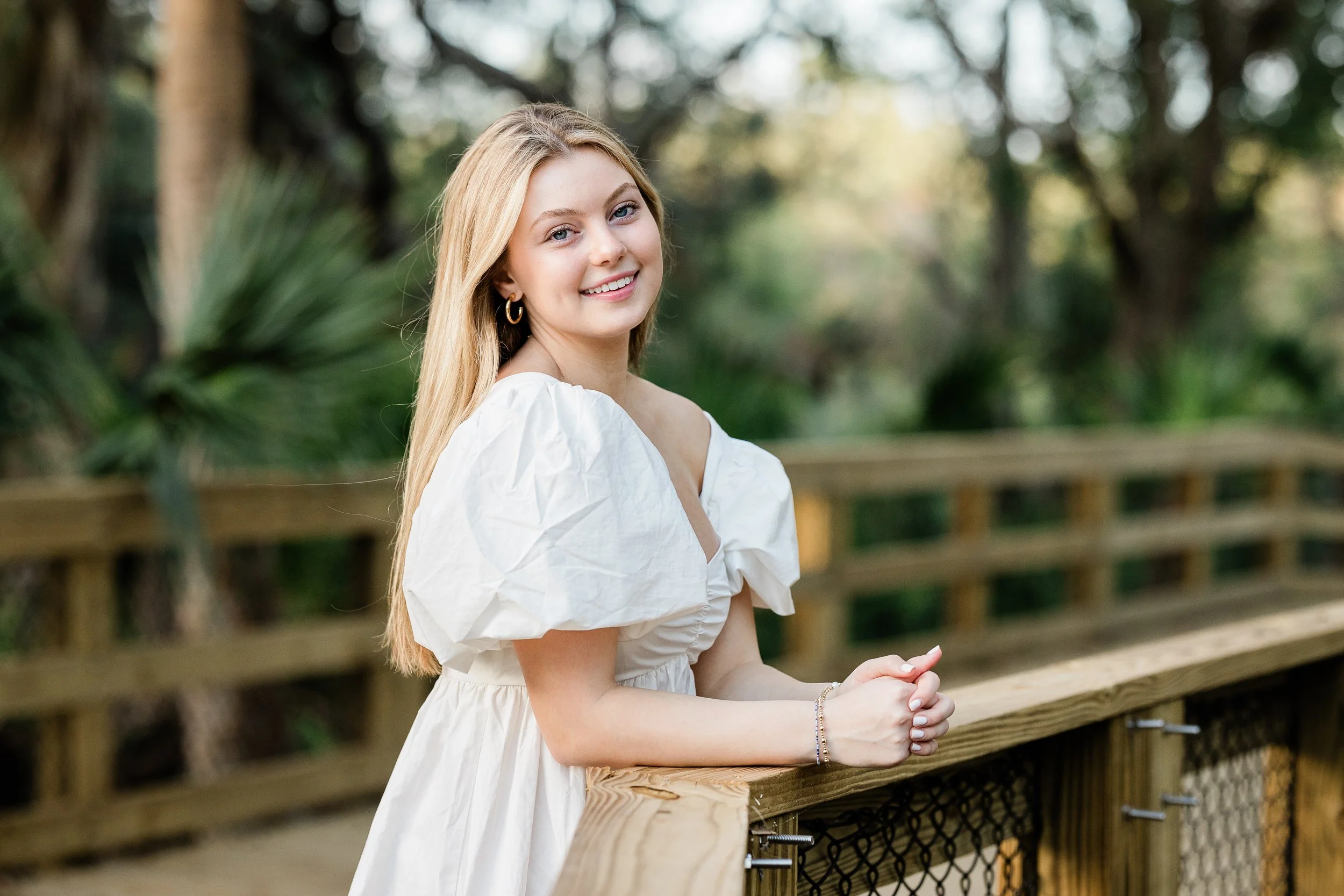 A young woman with long blonde hair smiling and standing outdoors by a wooden railing in a lush, green park or natural setting.