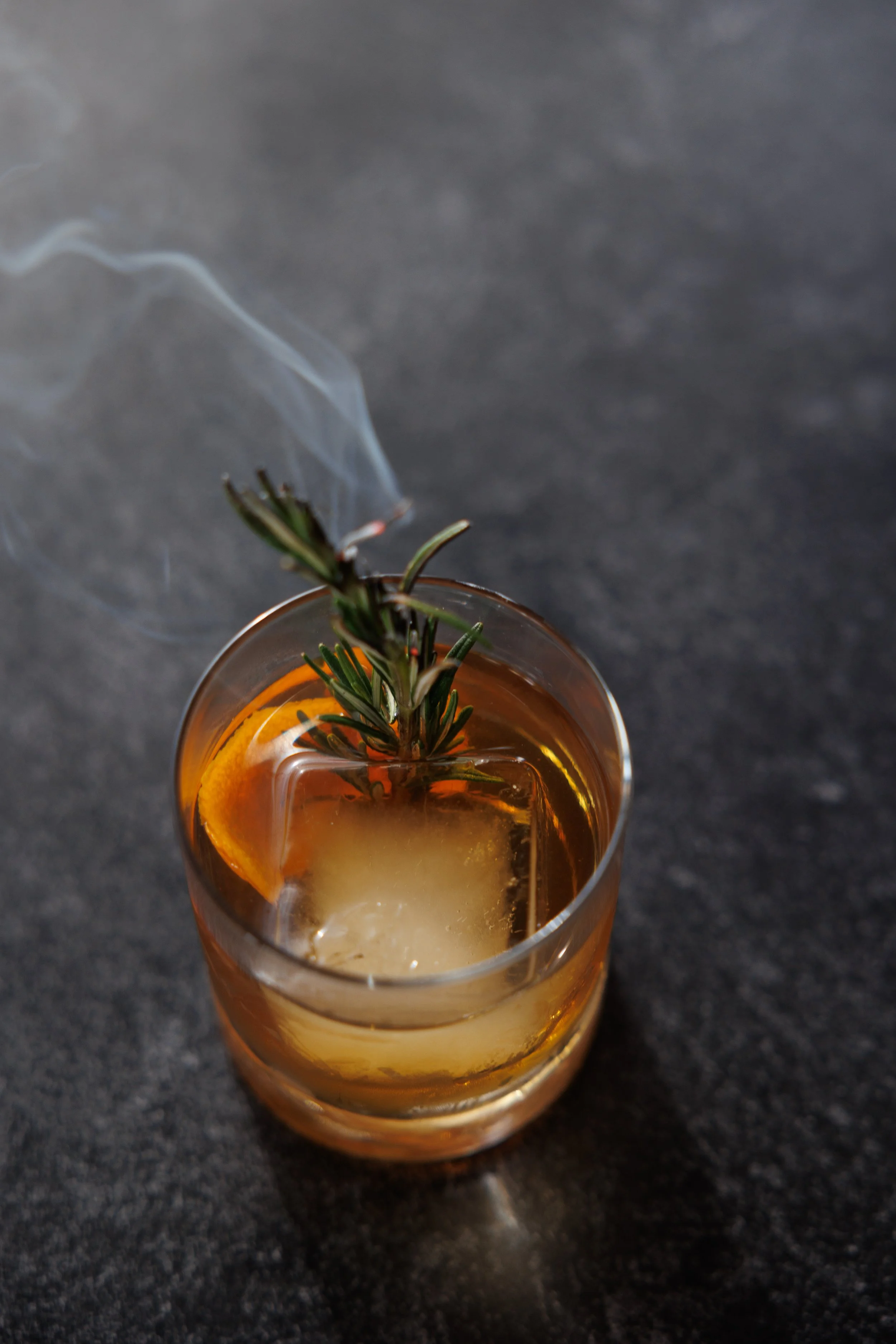 A glass of amber-colored beverage garnished with a slice of orange and a sprig of rosemary, with steam drifting upward.