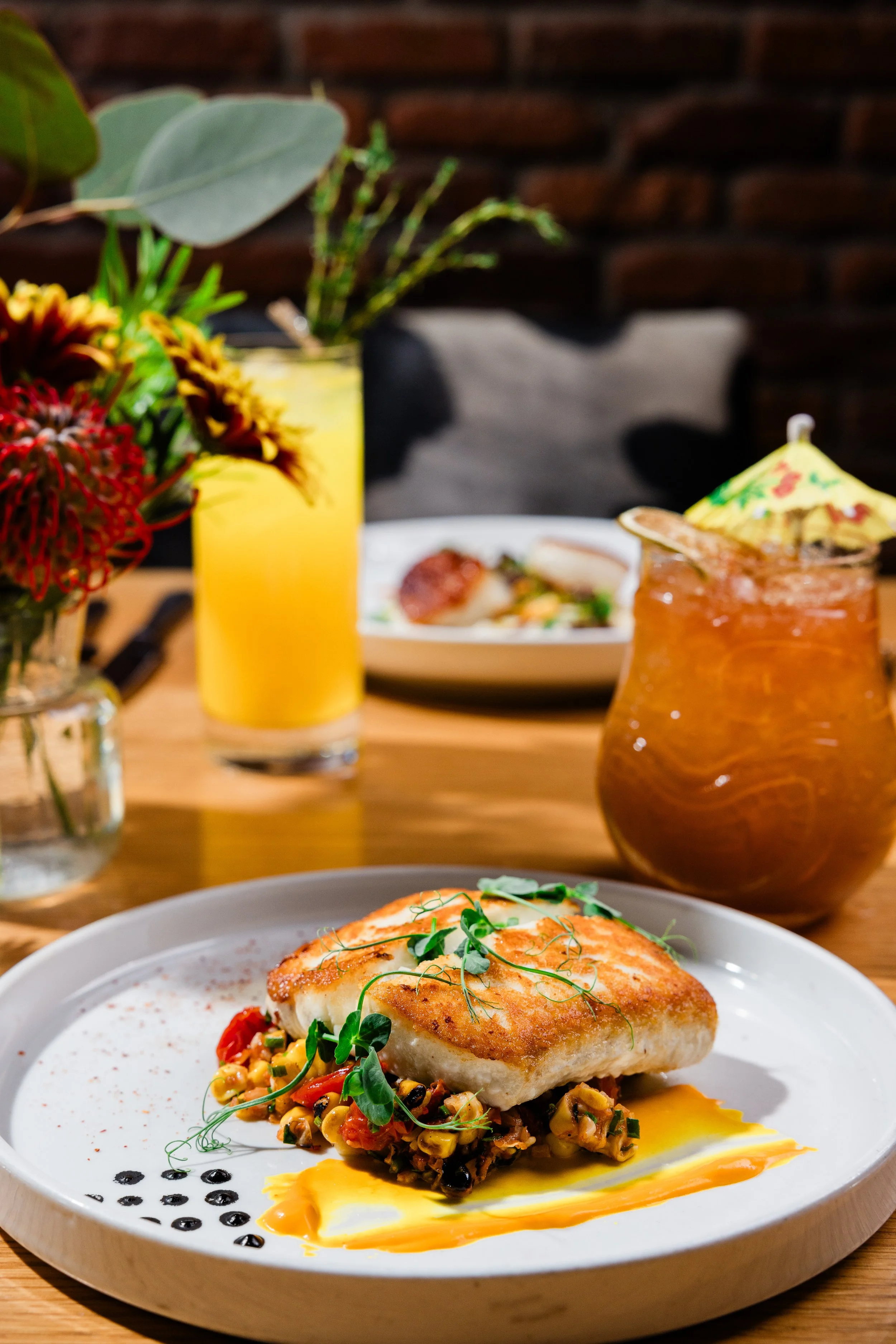 A plate of baked fish with a sauce, garnished with microgreens, served over a bed of vegetables and corn, with a drizzle of sauce on the plate. In the background, there is a glass of orange juice, a vase with flowers, and a cocktail with a paper umbr