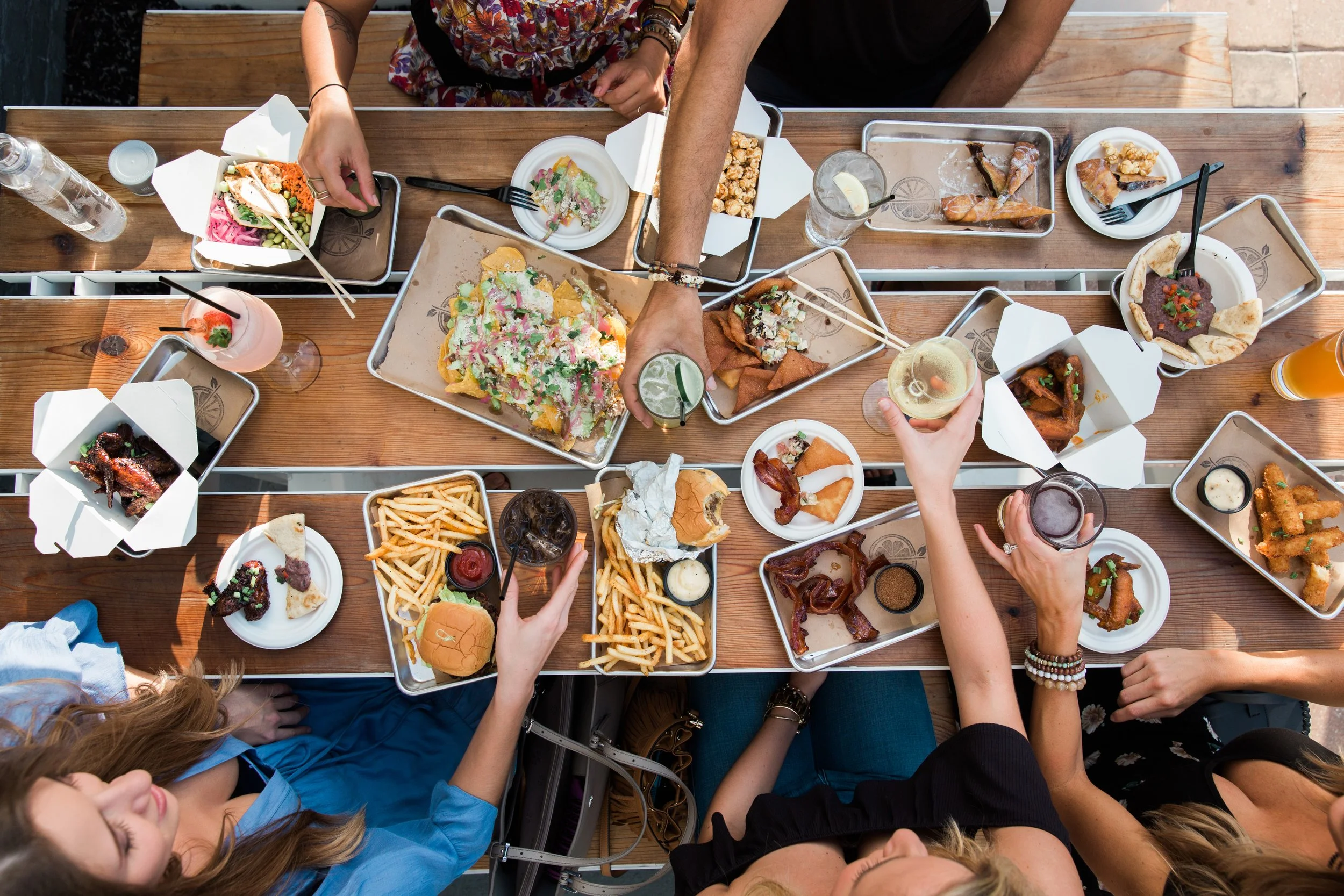 Top-down view of a wooden table filled with various dishes, drinks, and people reaching for food at an outdoor gathering.