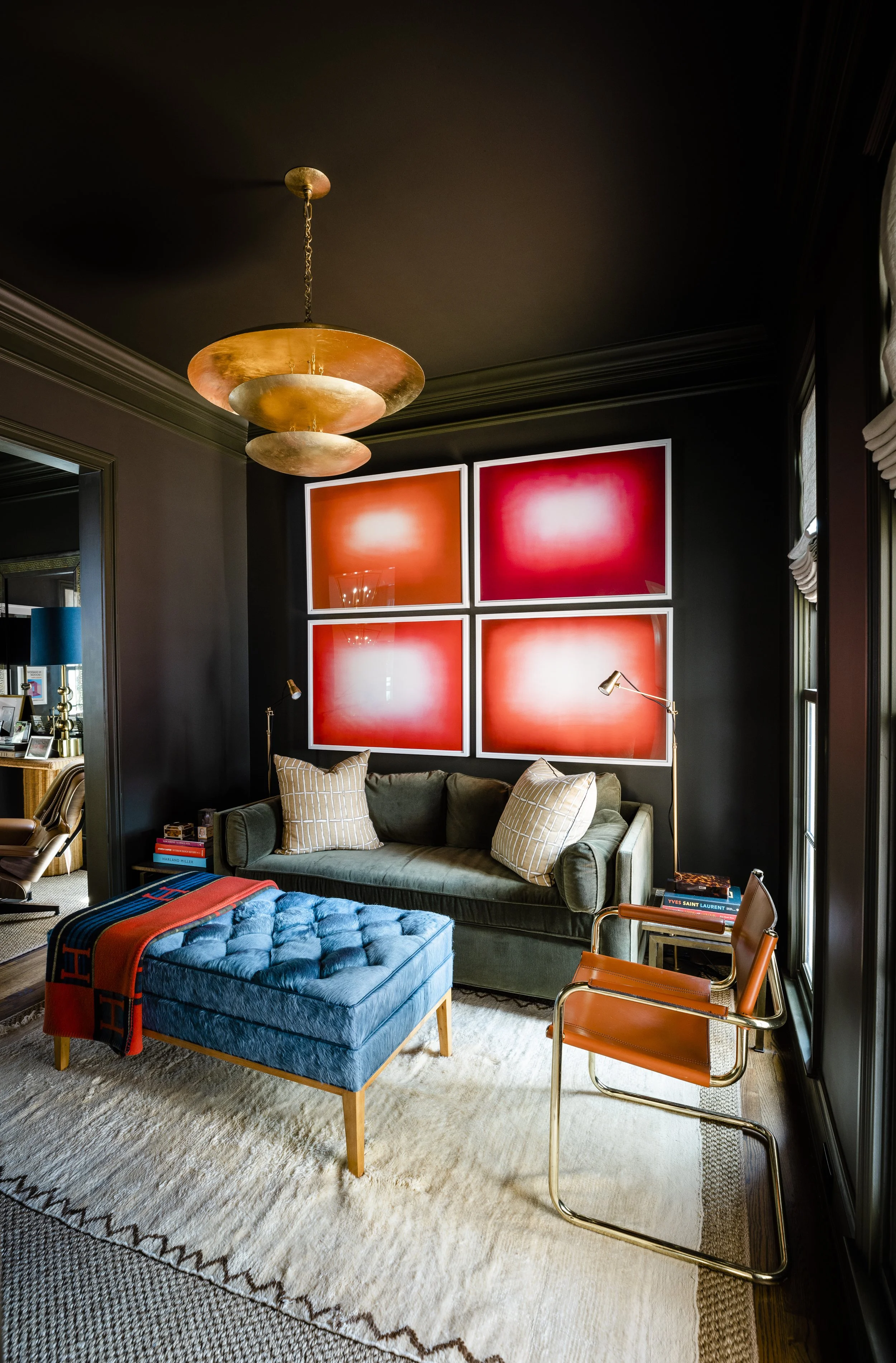 Living room with dark walls, a green velvet sofa with beige and white striped pillows, a blue tufted ottoman with a red scarf, modern orange and metallic chairs, wall art with four large red squares, a chandelier with golden and copper discs, a side 