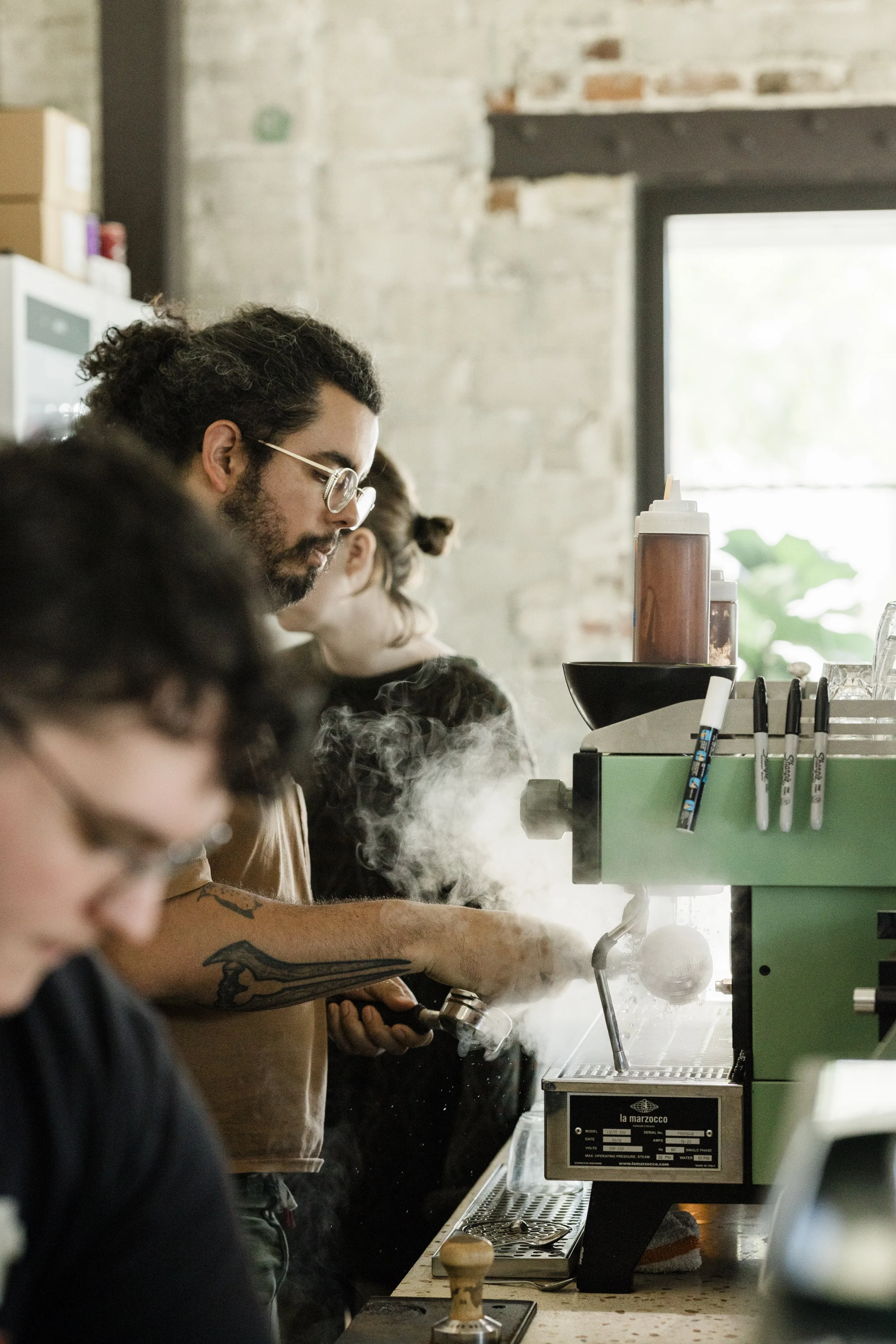 People standing in a line at a coffee shop counter, with various drink-making equipment, including a squeeze bottle and a coffee machine, in a cafe with brick walls and a window in the background.