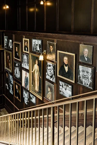 Gallery wall with framed black and white and color portraits and photographs, above a staircase railing in a dimly lit room.