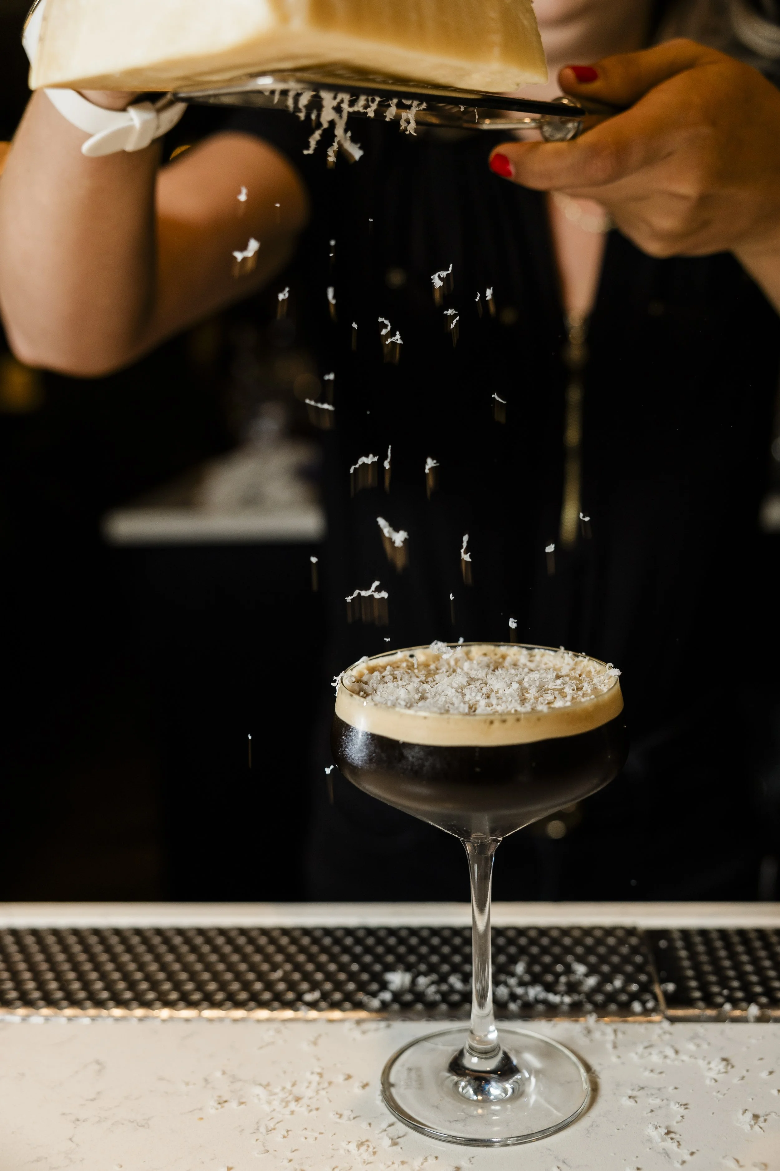 A cocktail glass filled with a dark beverage and topped with shredded coconut, with a bartender grating coconut over the drink.