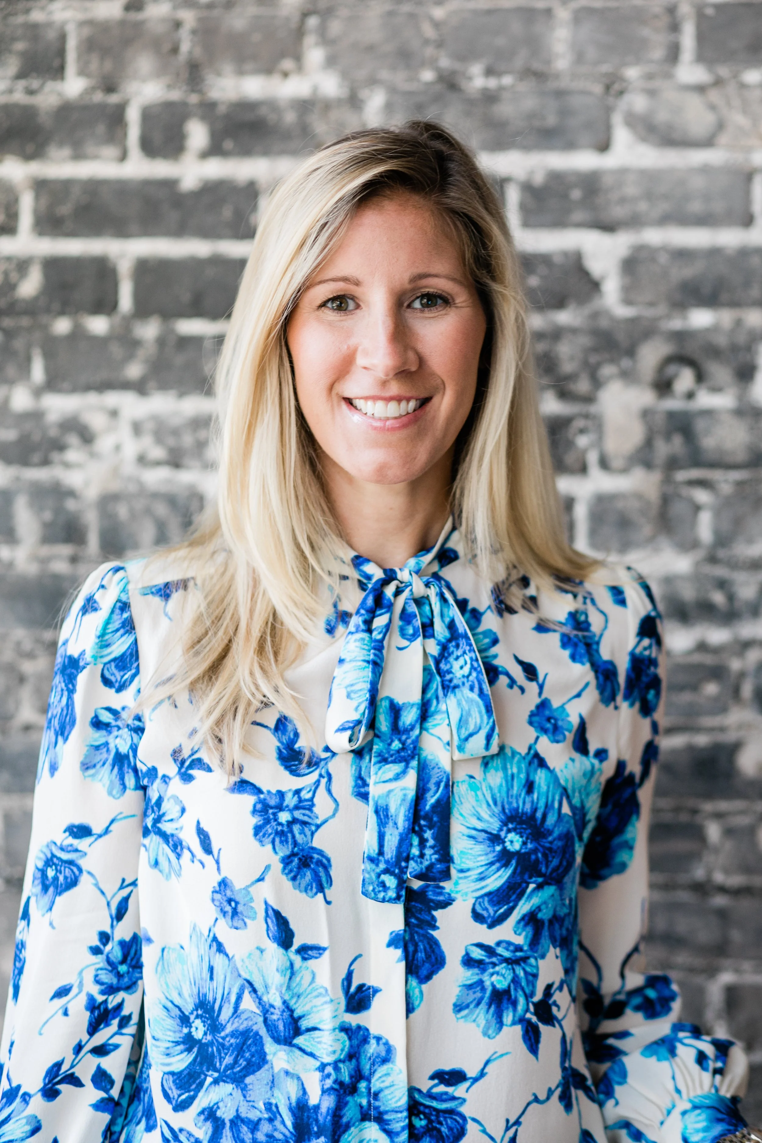 indoor environmental portrait of a woman with blond hair and wearing a blouse with blue flowers, background is grey brick