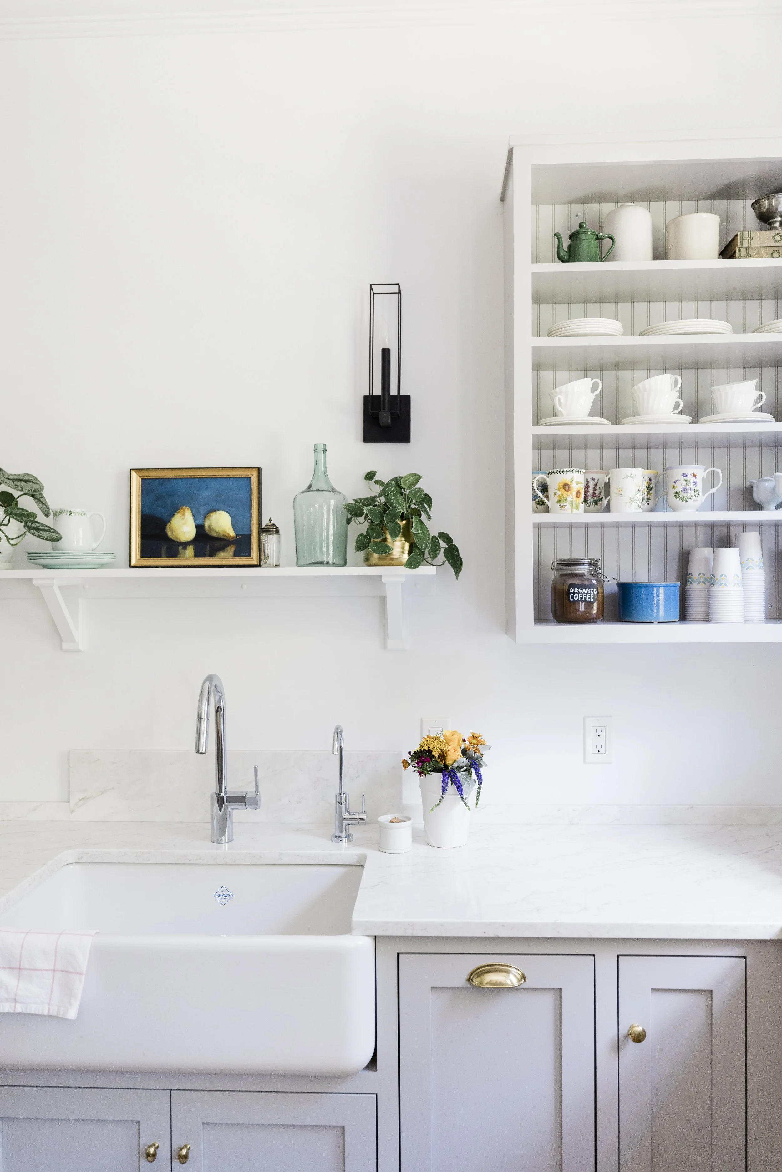 A white kitchen with a farmhouse sink, gray cabinets with gold knobs, and open shelving displaying dishes, cups, and a green teapot. There is a vase with flowers on the countertop. Interior design by jamie finley & co.