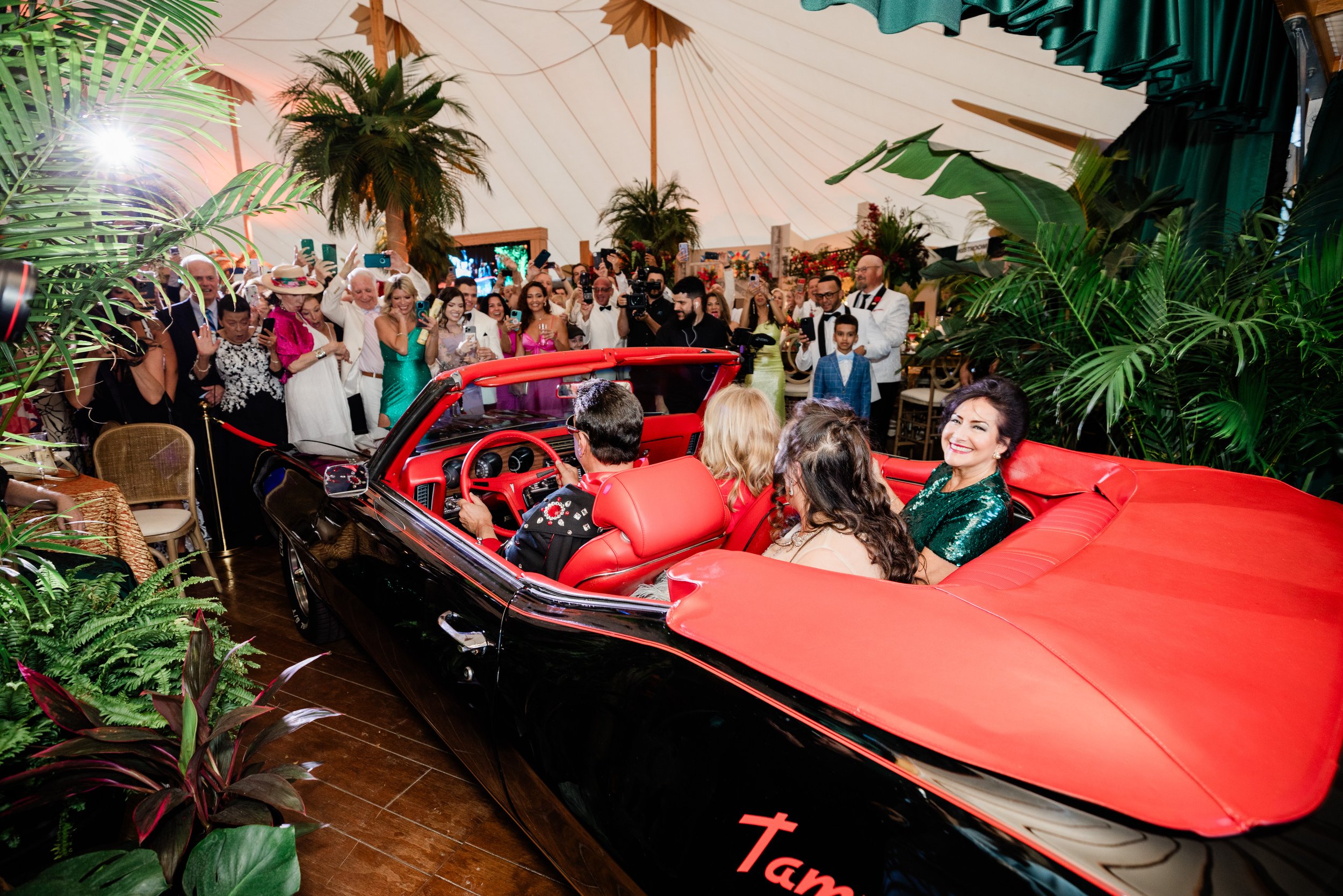 A large gathering of people dressed in formal attire inside a decorated tent, with some taking photos or recording videos. There is a vintage red and black convertible car in the foreground, surrounded by lush green plants.