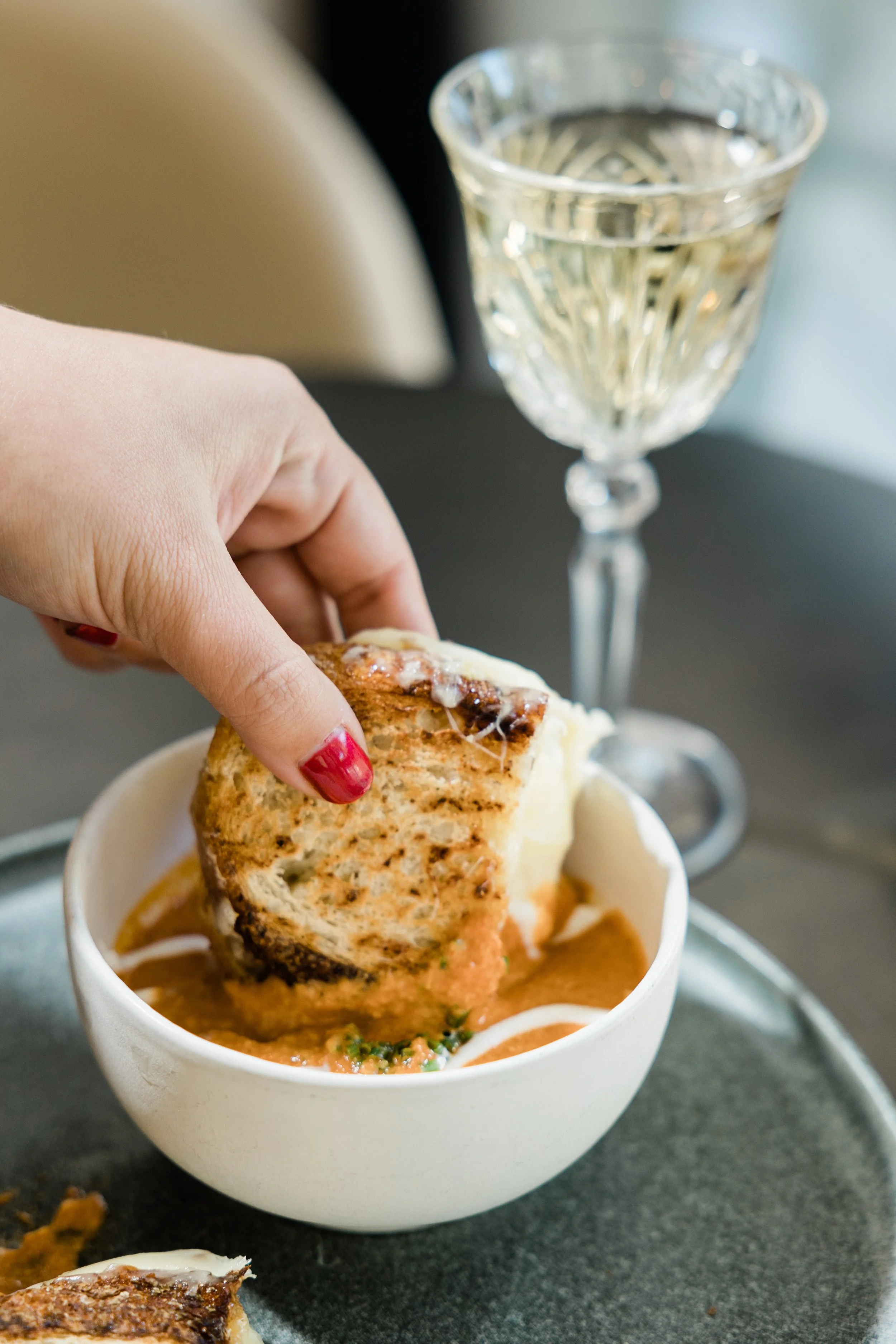 A person with red nail polish dips a piece of naan bread into a bowl of curry with a glass of white wine in the background.