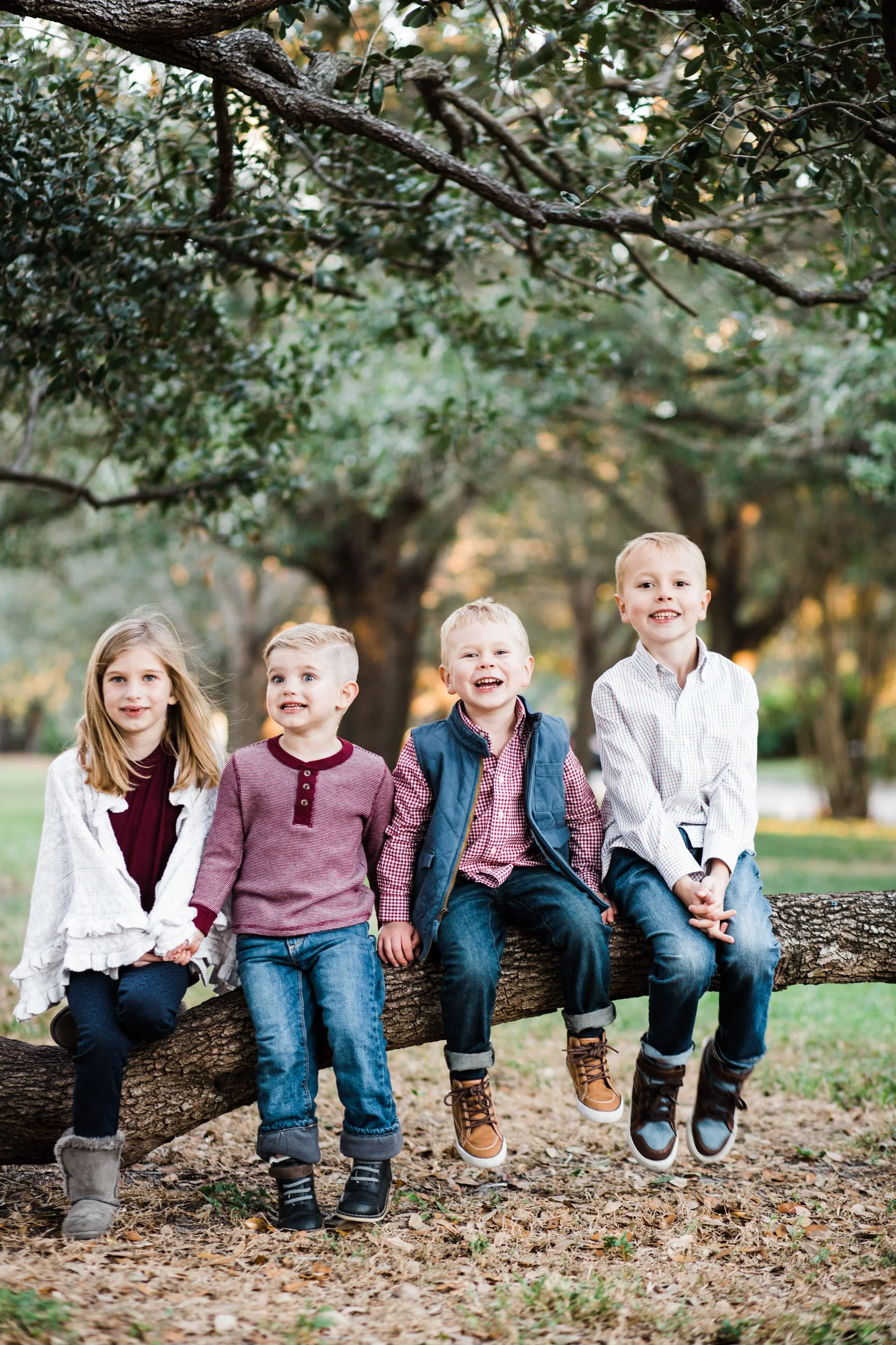 Five children sitting on a tree branch outdoors in a park, smiling and looking at the camera, with green trees and fallen leaves in the background.