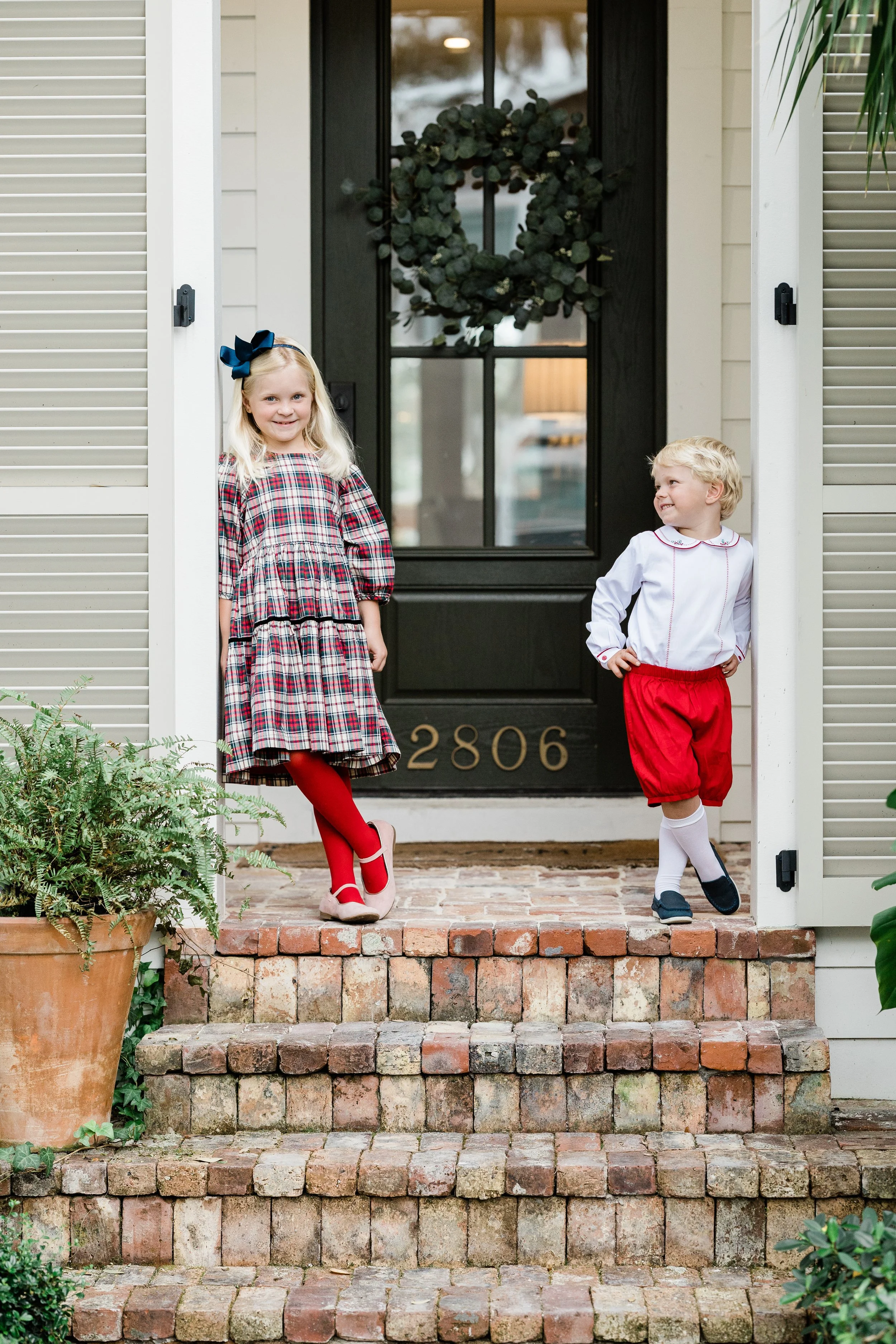 A young girl and boy stand on brick steps in front of a house, smiling. The girl wears a plaid dress, red tights, beige shoes, and a large navy bow in her hair. The boy wears a white shirt with red piping, red shorts, and navy shoes. Behind them, a b