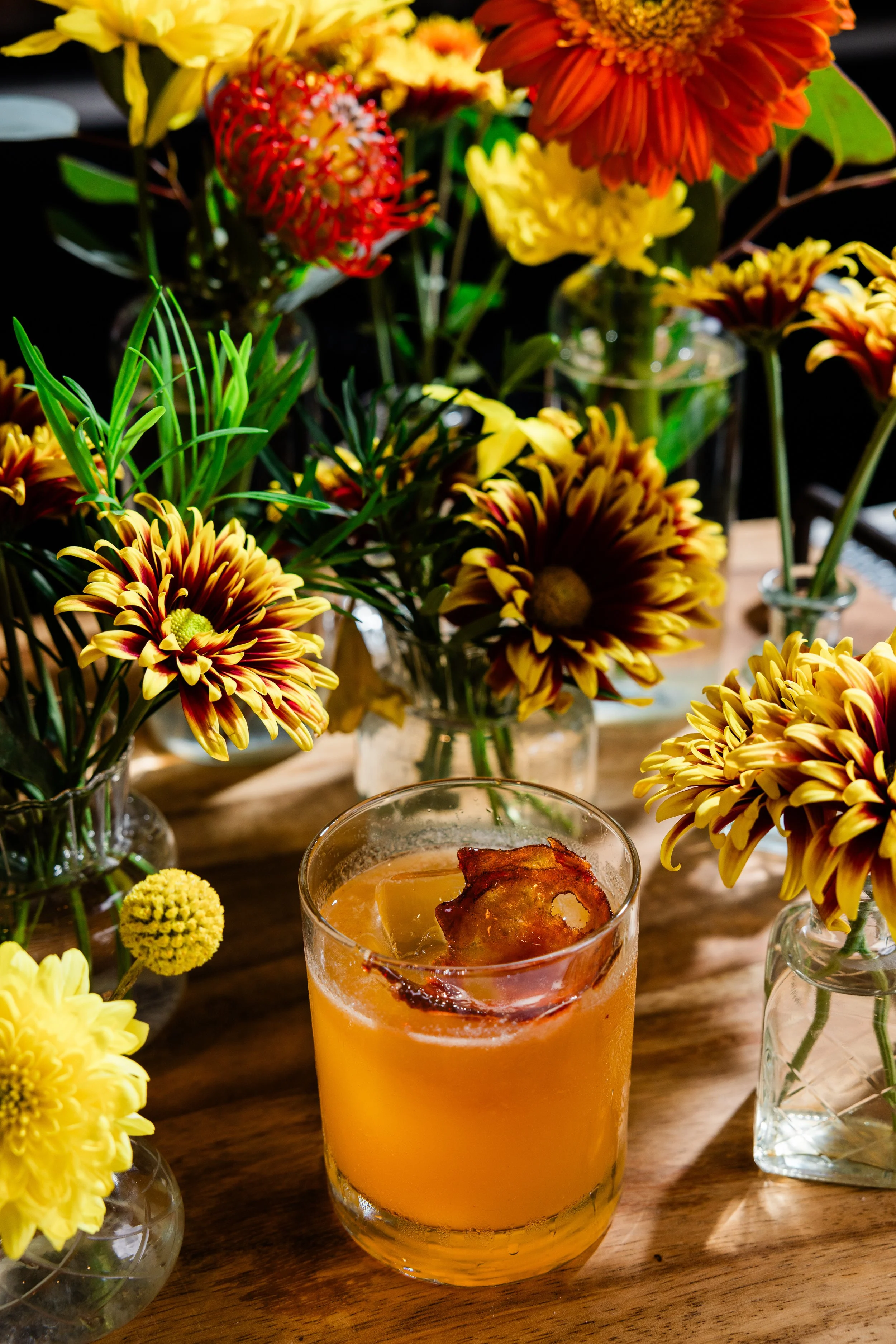 A close-up of a fruit cocktail with ice and bacon in a glass, surrounded by various colorful flowers in vases on a wooden table.