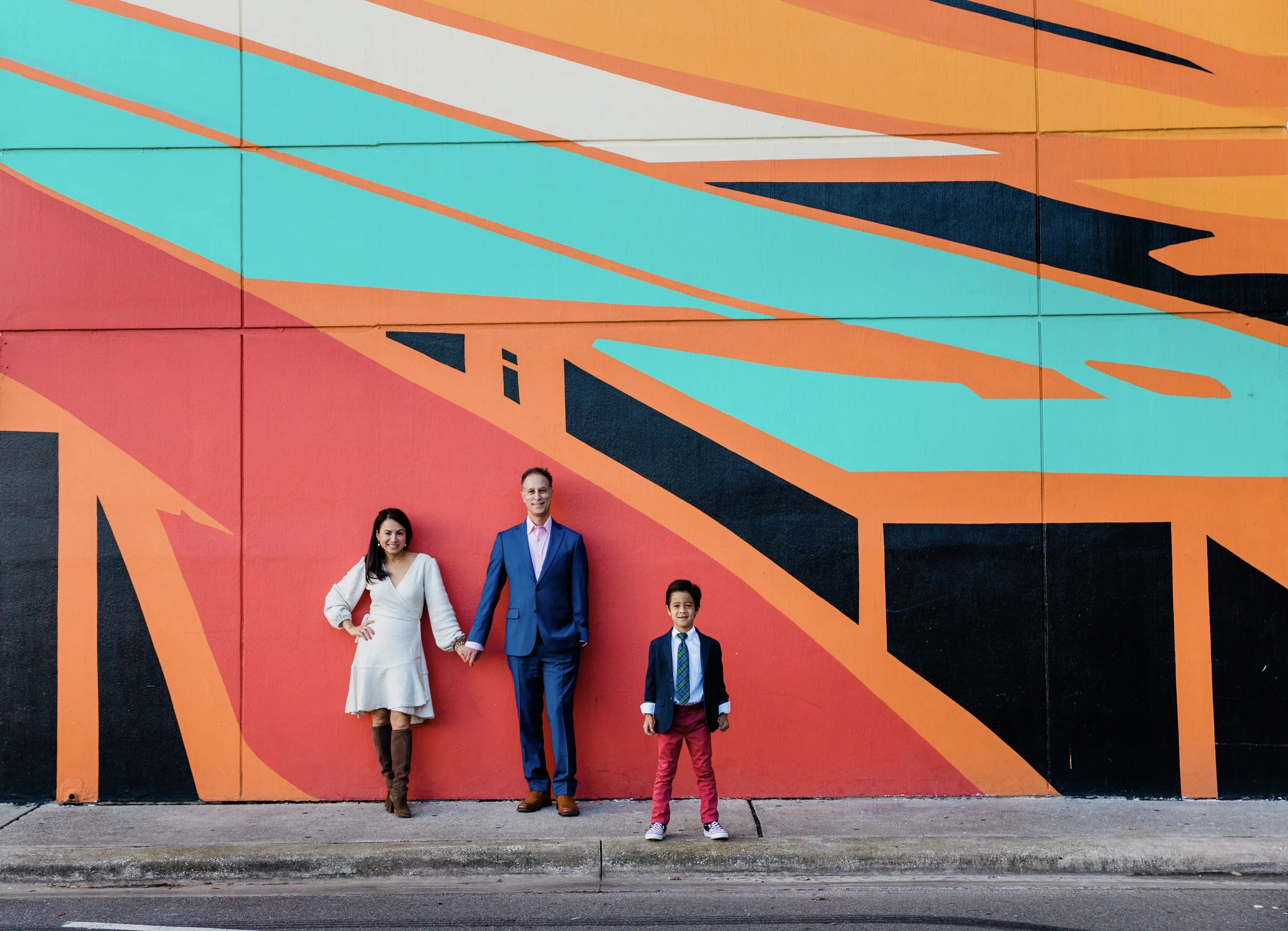 woman in a white sweater dress, man in a blue suit, young boy in red pants and a jacket and tie in front of a dramatic red, orange, teal, and black mural in downtown Tampa.
