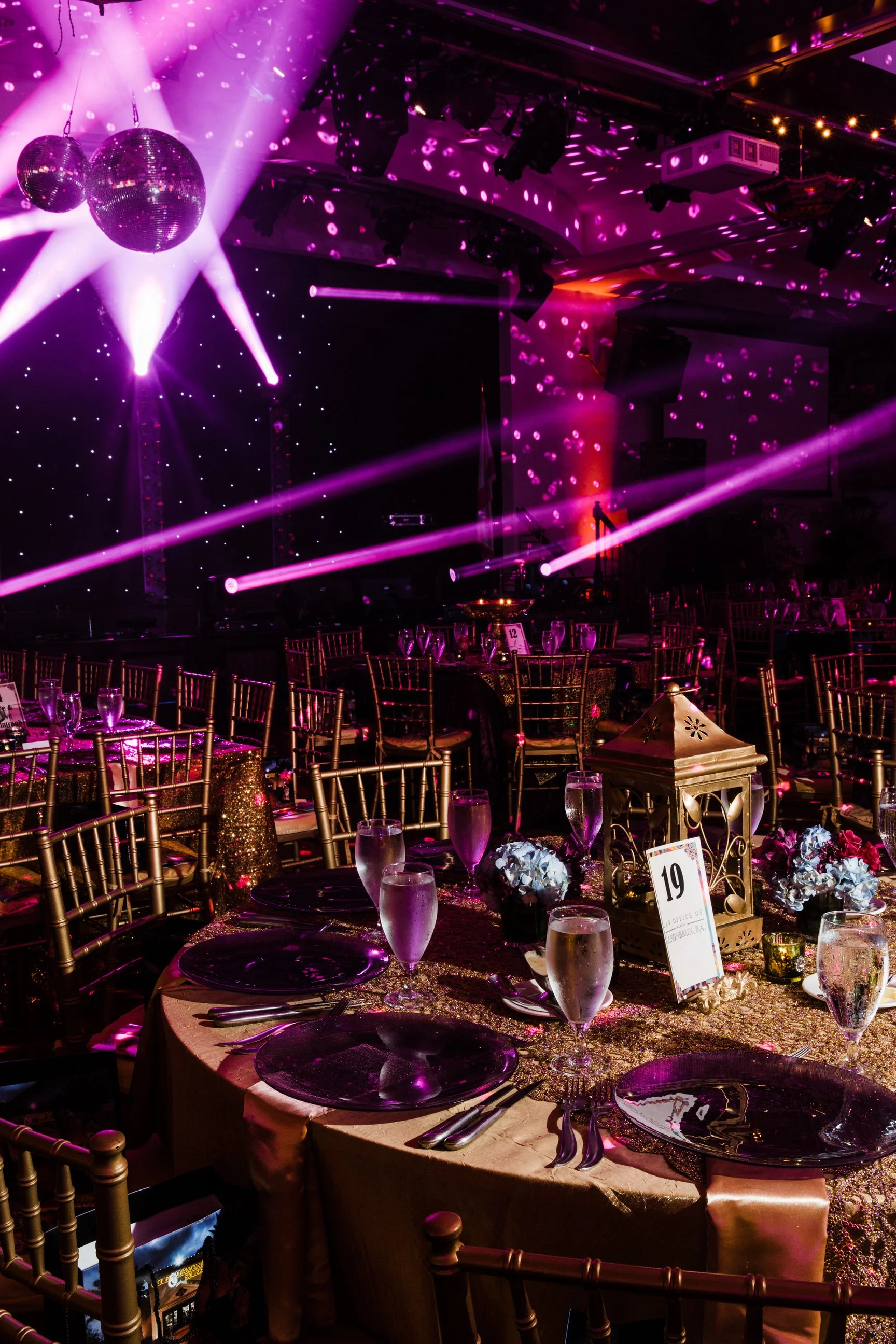 A banquet hall decorated for a celebration with round tables, gold chairs, flower centerpieces, and a glittering gold tablecloth. The room is illuminated with pink and purple spotlights, and disco balls hang from the ceiling, reflecting light across 